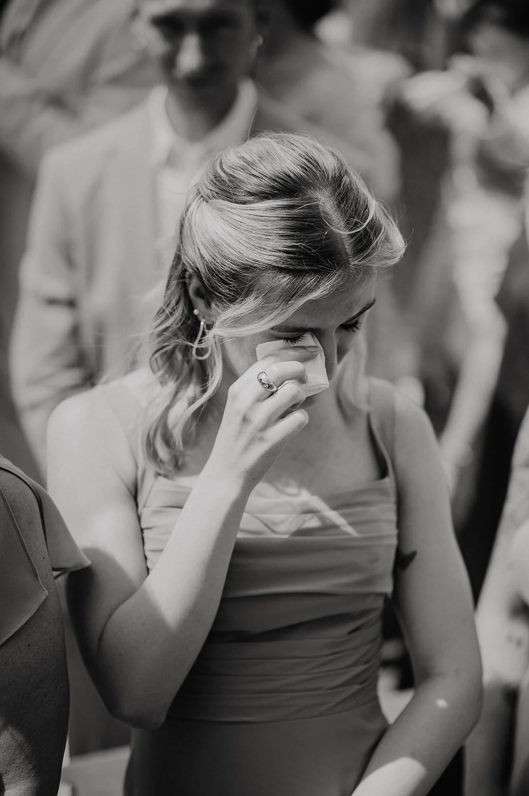 A young woman with styled hair is crying and wiping her eyes with a tissue during a emotional moment, with a man standing behind her in a crowded setting.