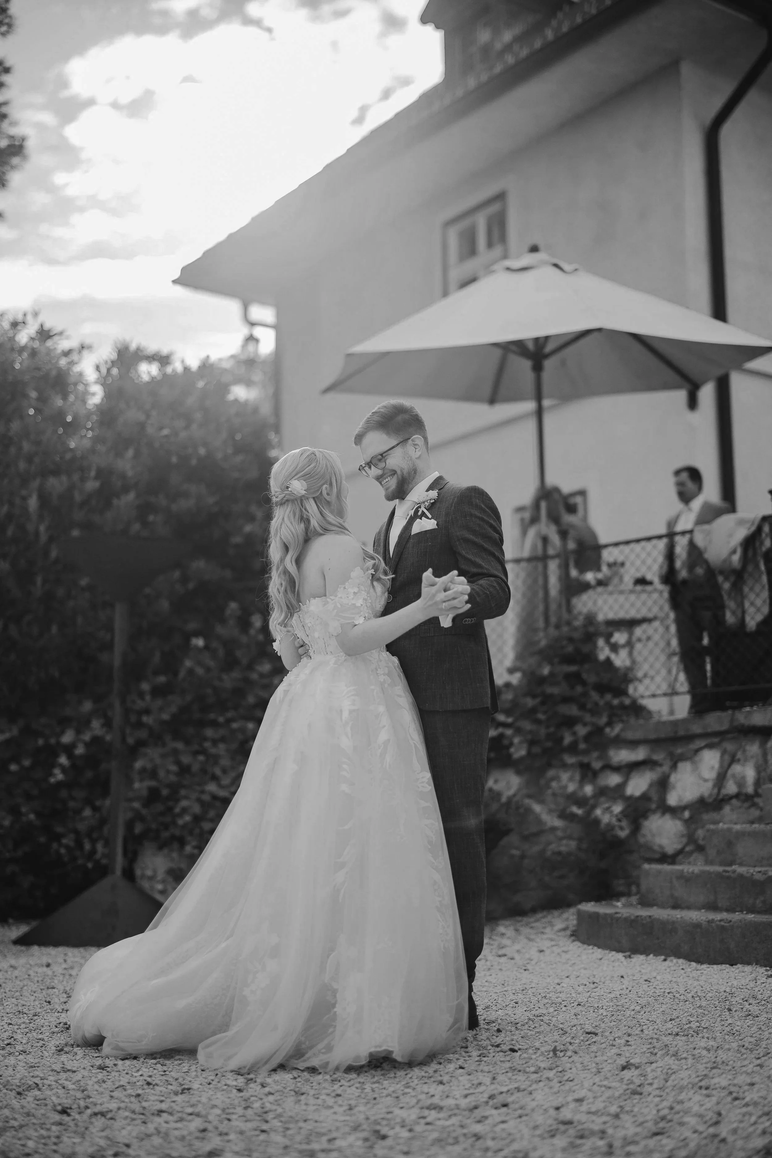 Ein Brautpaar tanzt während seiner Hochzeit im Freien. Die Braut trägt ein langes Spitzenkleid mit schulterfreiem Ausschnitt, und ihr Haar ist mit einem Blumenaccessoire verziert. Der Bräutigam trägt einen dunklen Anzug und eine Brille. Sie lächeln und schauen sich an, im Hintergrund sind ein großer Regenschirm und ein Haus zu sehen.