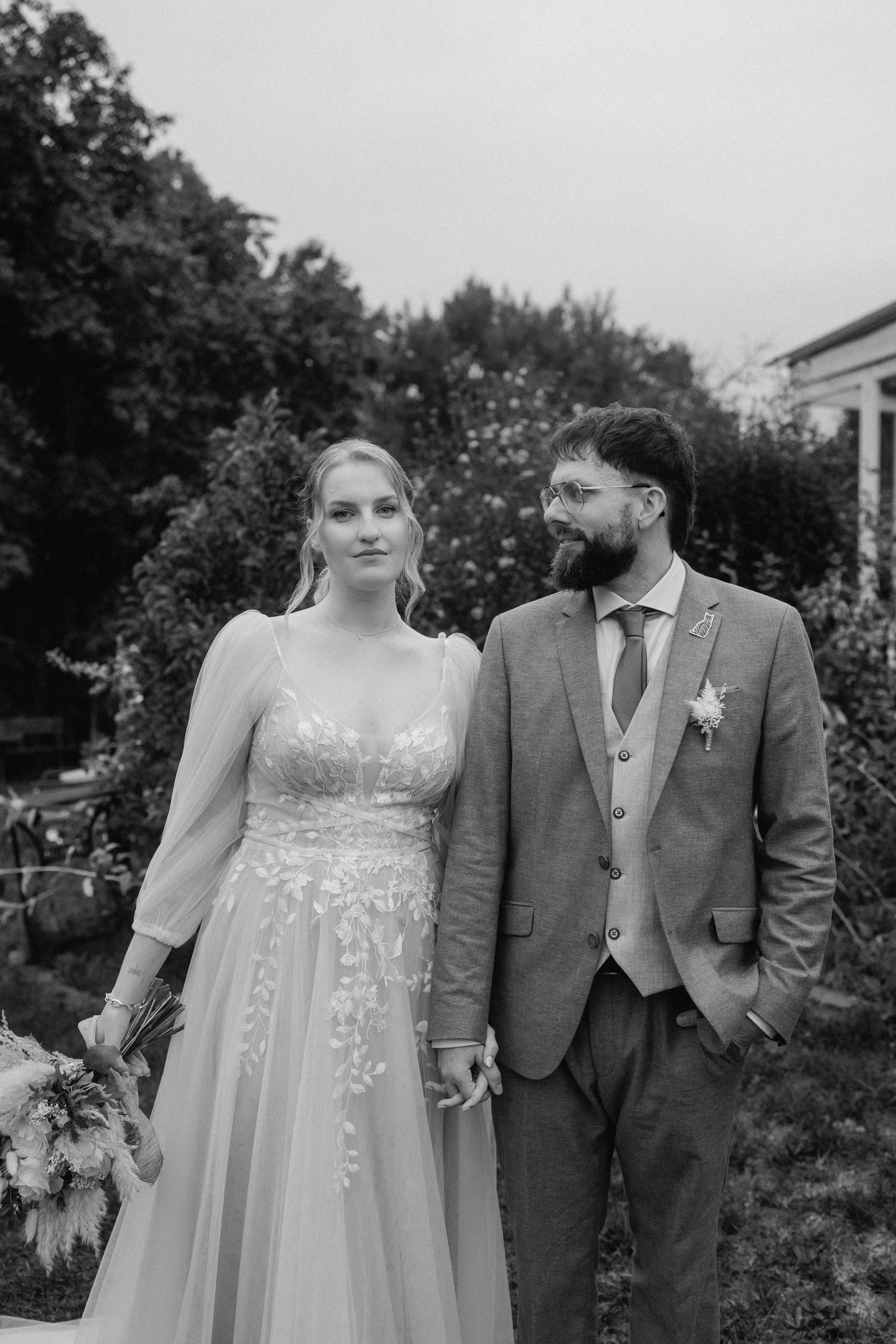 Ein Schwarz-Weiß-Foto eines Brautpaares, das im Freien steht und Händchen hält. Die Braut trägt ein Brautkleid mit Spitzenverzierungen und hält einen Blumenstrauß in der Hand, während der Bräutigam einen Anzug mit Ansteckblume und eine Brille trägt. Sie sind von Bäumen umgeben, und im Hintergrund ist ein Haus zu sehen.