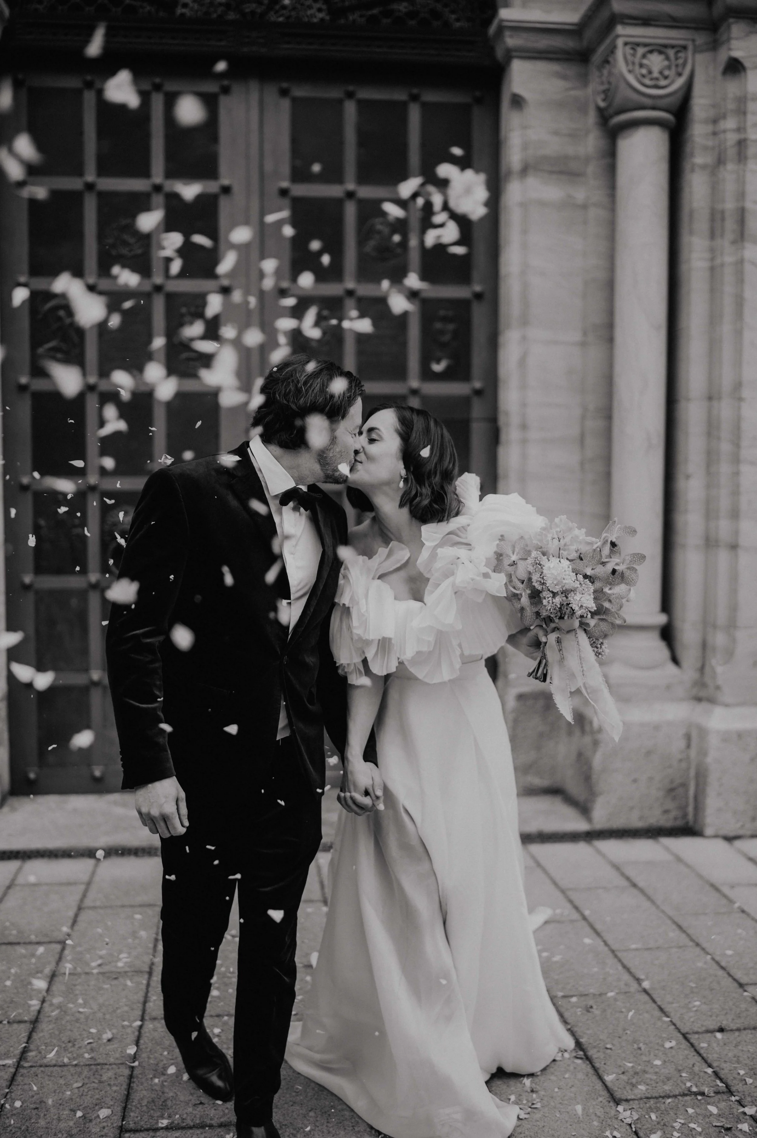 Ein Schwarz-Weiß-Foto eines frischvermählten Paares, das sich nach der Hochzeit küsst, Händchen hält und von herabfallenden Blütenblättern umgeben ist, während es vor einem Gebäude mit kunstvoller Architektur und Säulen steht.