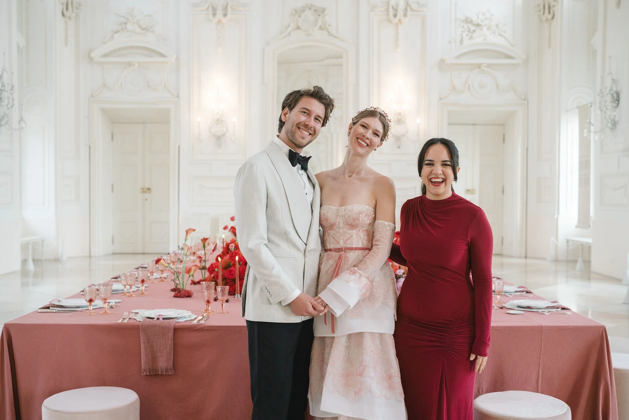 Ein lächelndes Hochzeitspaar und eine Frau stehen in einem eleganten, verschnörkelten weißen Raum mit einem dekorierten Esstisch im Hintergrund.