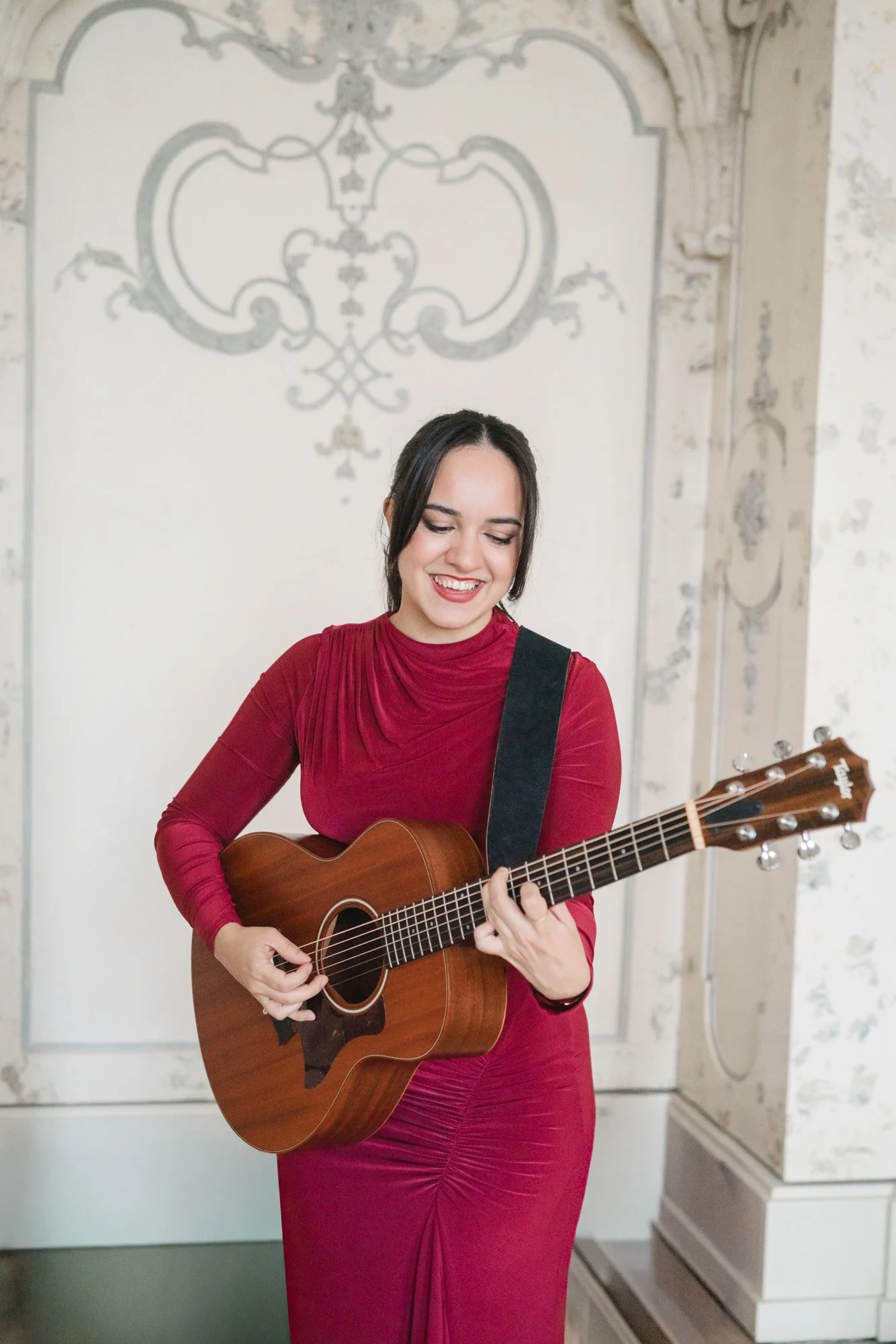 Eine Frau mit dunklem Haar und rotem Kleid spielt auf einer akustischen Gitarre und lächelt.