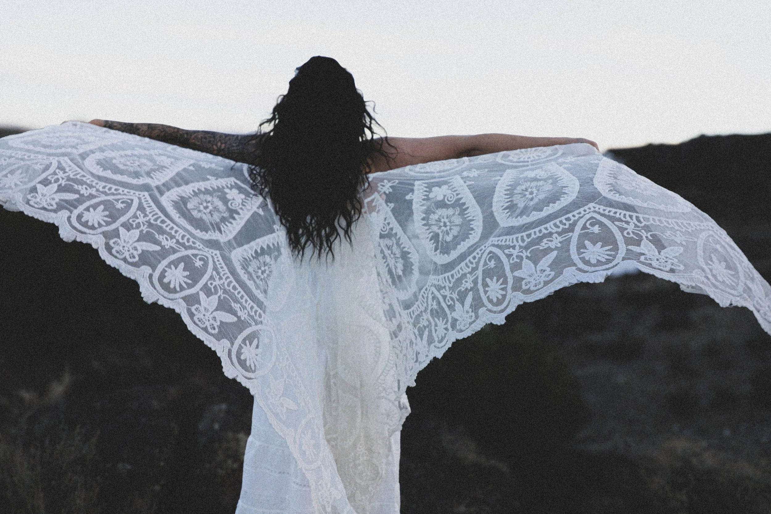 Woman seen from behind, arms outstretched in a white lace shawl like wings