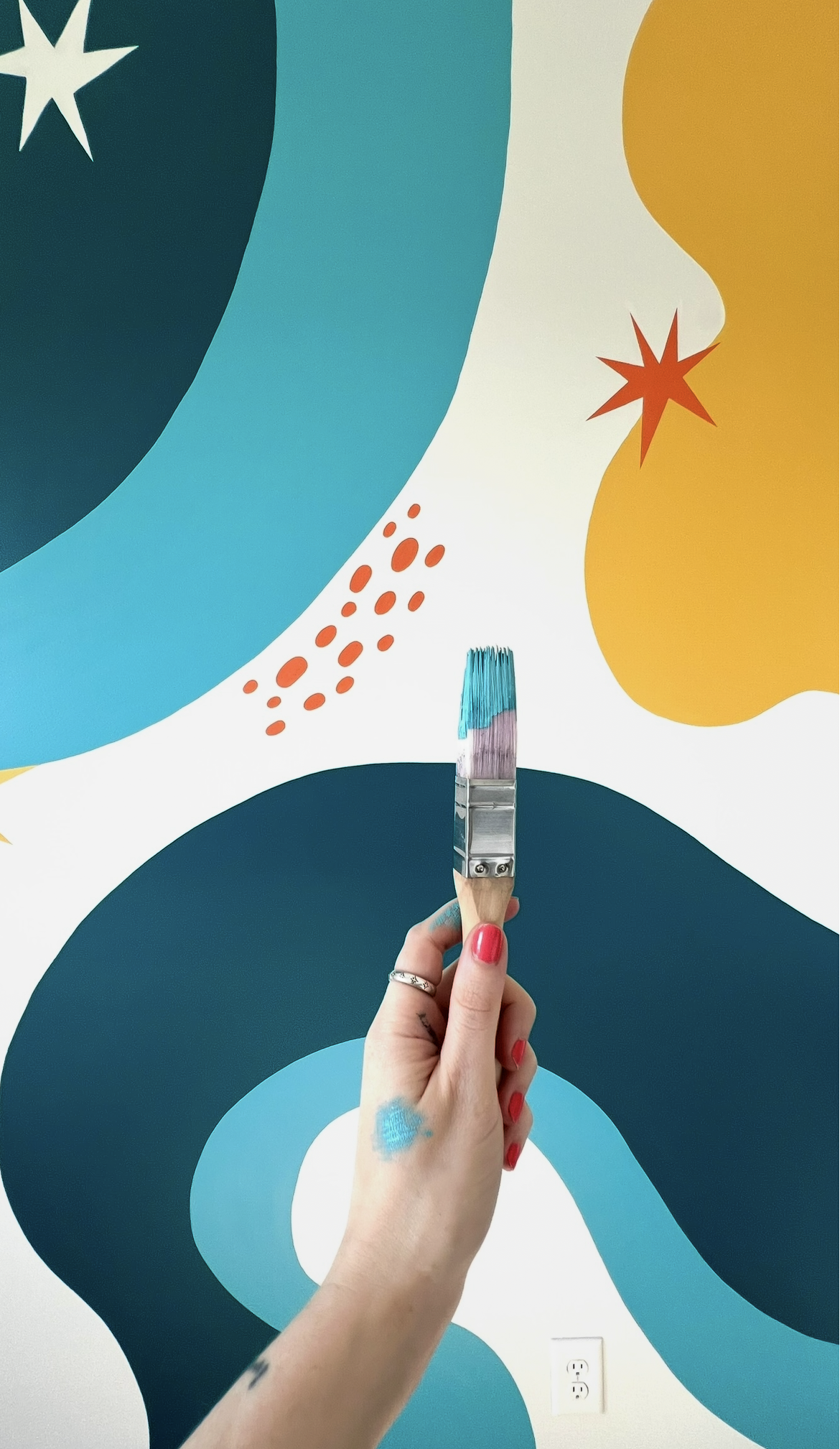 Person holding a paintbrush with blue paint on it, painting a colorful abstract mural with large curved shapes in shades of blue, yellow, and black, with red accents.