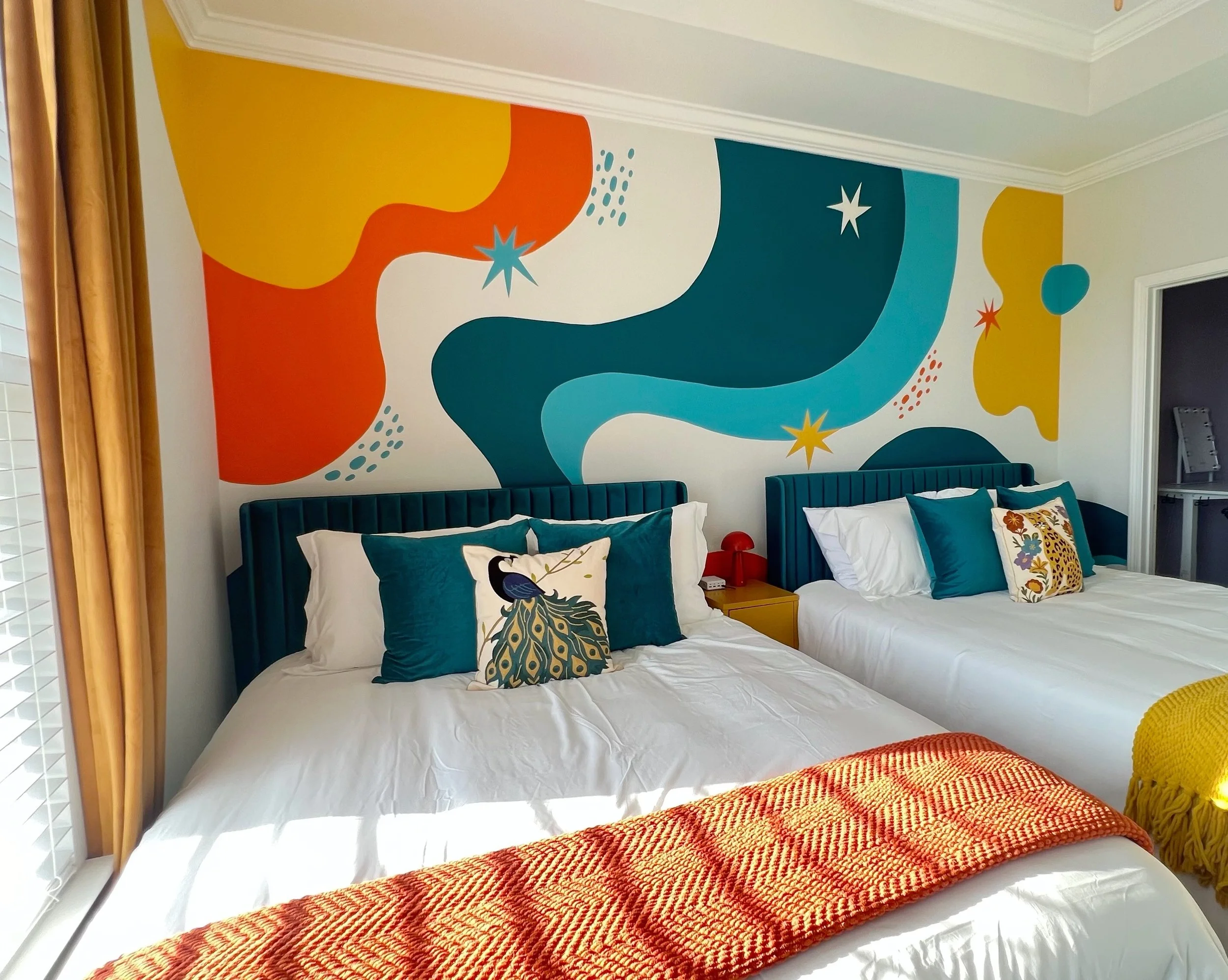 A bedroom with a colorful abstract mural on the wall behind the beds, featuring flowing shapes in yellow, orange, blue, and teal with starburst patterns. Two beds with teal velvet headboards are in the foreground, decorated with pillows including one