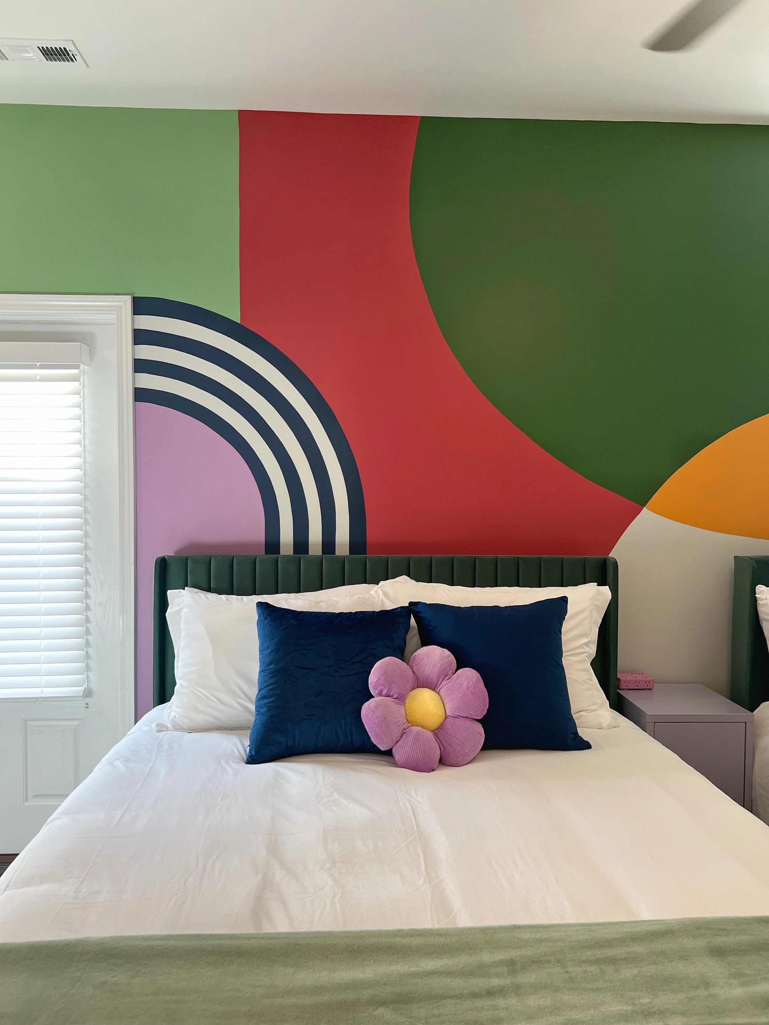 A neatly made bed with white sheets, two dark blue pillows, and a pink flower-shaped pillow with a yellow center. The bed is against a colorful abstract painted wall with green, red, purple, and yellow segments. There is a window with white blinds on