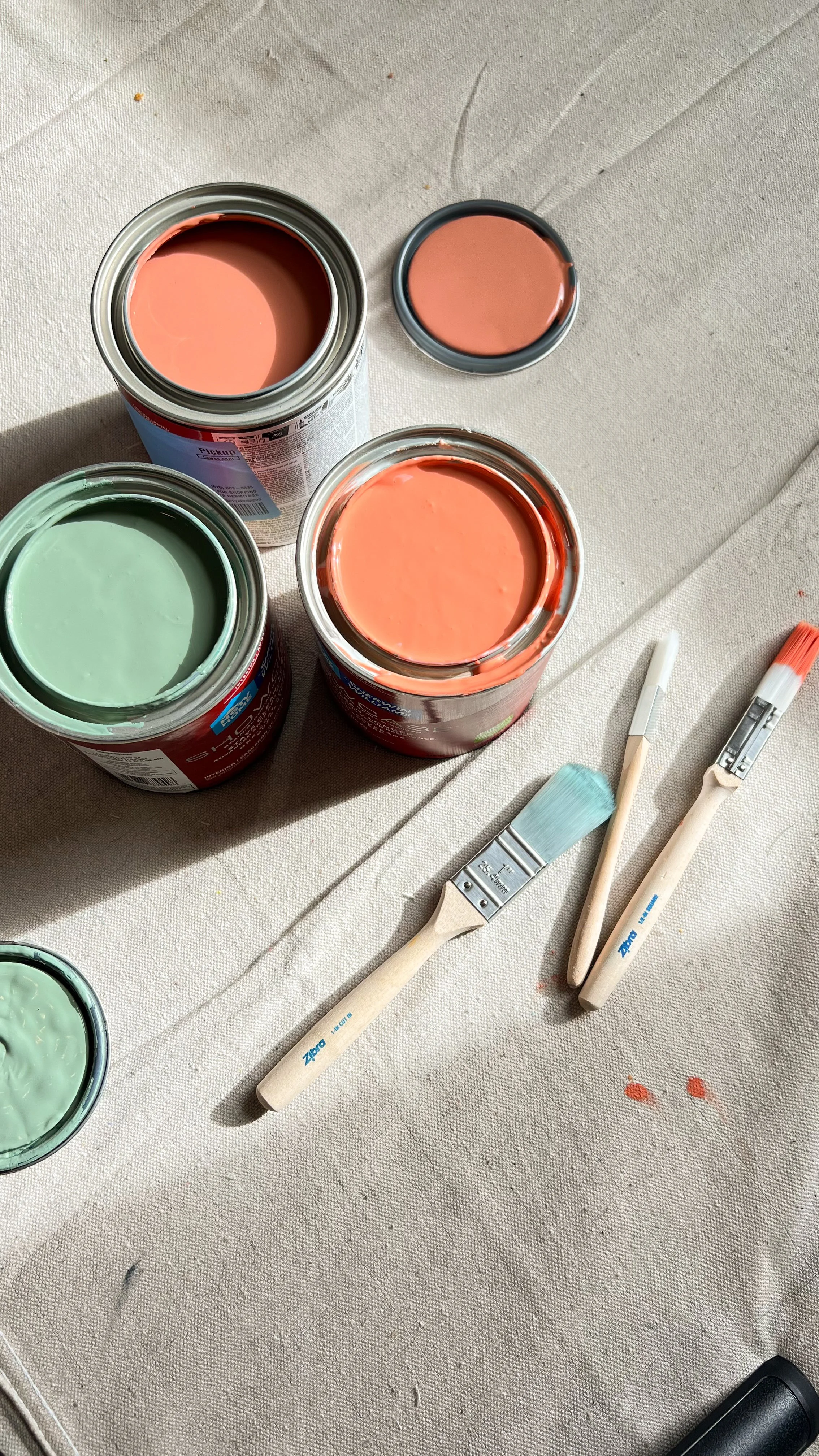 Open paint cans with pink and green paint, paintbrushes and a spatula on a beige surface.