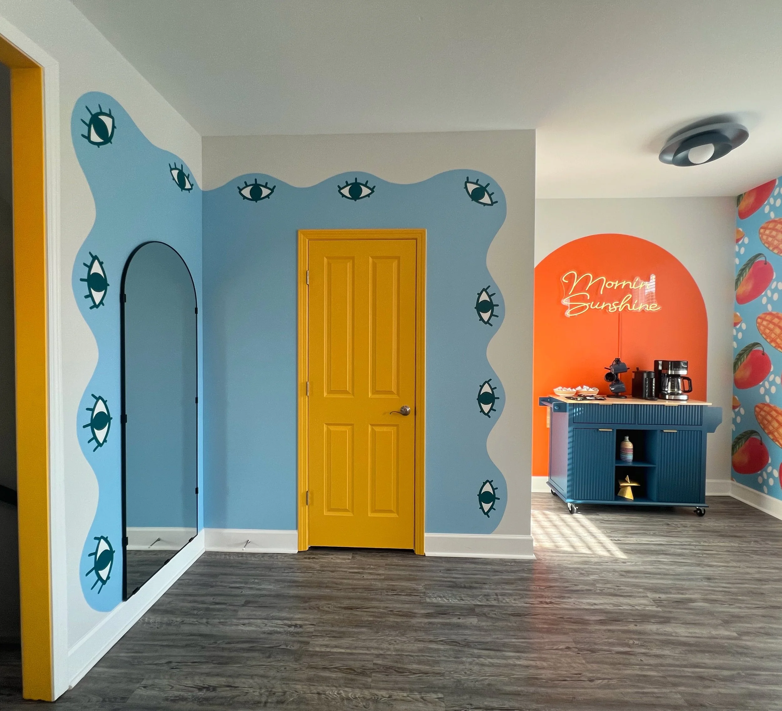 Interior of a colorful room with walls painted half white and half blue, decorated with black eye illustrations, a yellow door, a small blue rolling cart with kitchen appliances, and a vibrant mural with red, orange, and yellow accents.