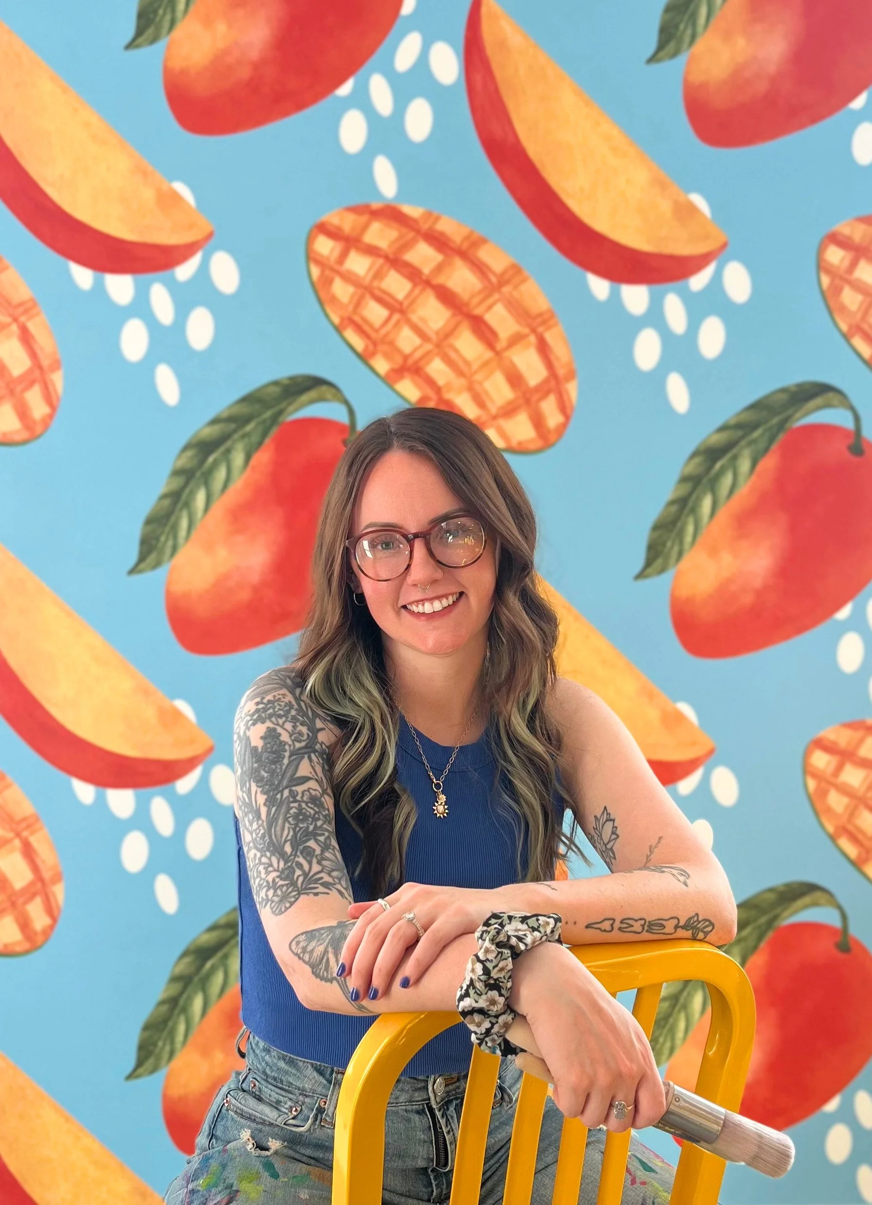 A woman with glasses, tattoos, and colorful hair smiling, leaning on a yellow chair, in front of a bright blue background with large, stylized peaches and peach slices.
