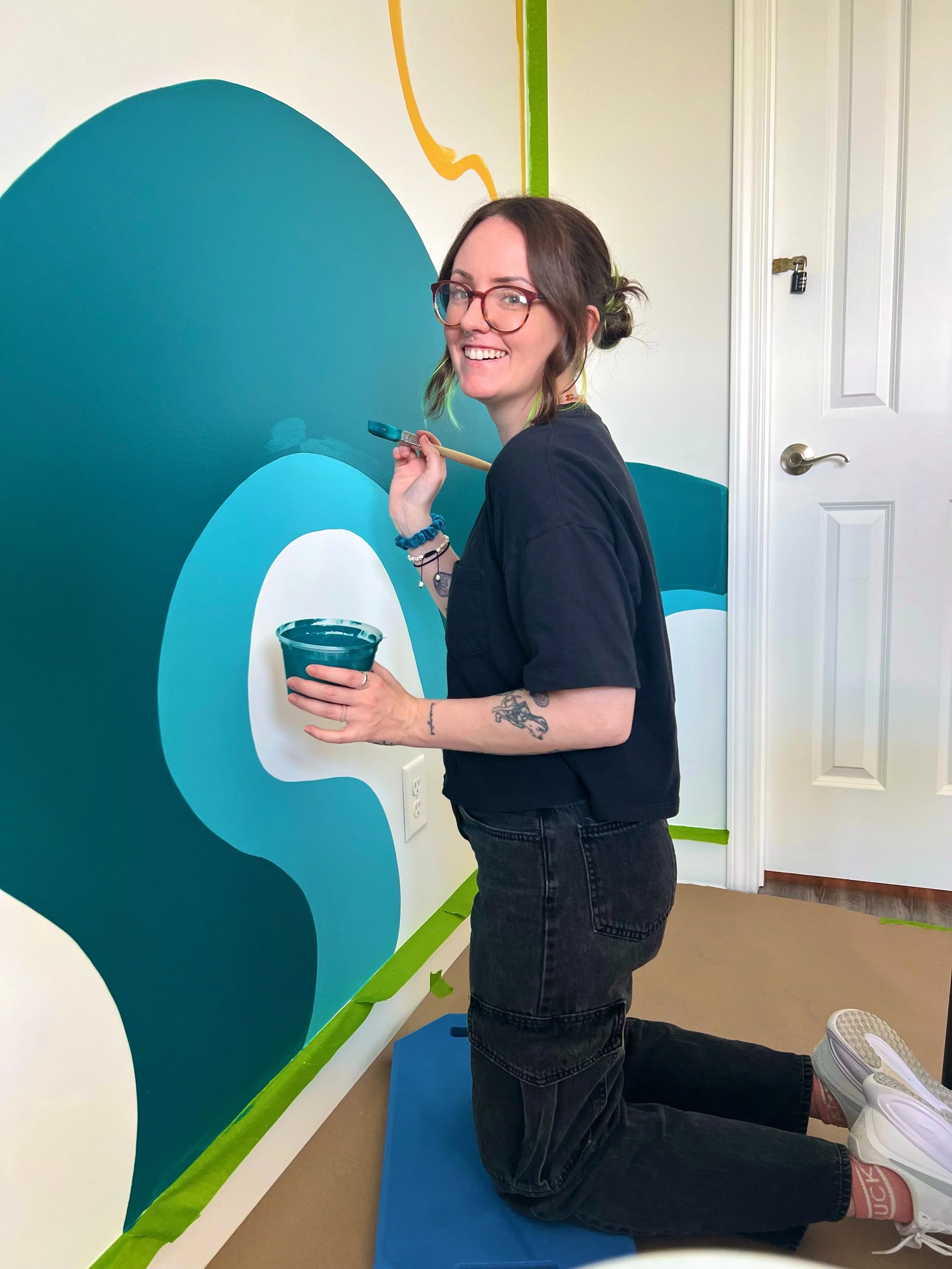 A woman with glasses and tattoos kneels on a mat, painting a wall with a paintbrush in a room decorated with colorful, abstract designs.