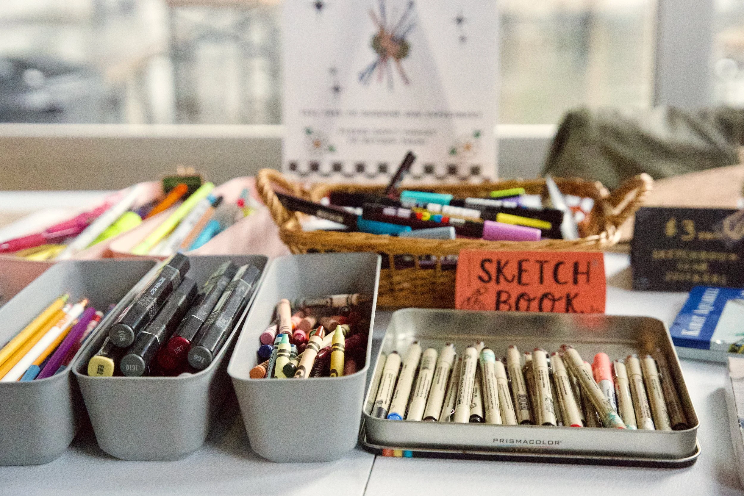 Containers filled with various colored markers, crayons, and pens on a table, with a sign labeled 'Sketch & Draw' in the background.