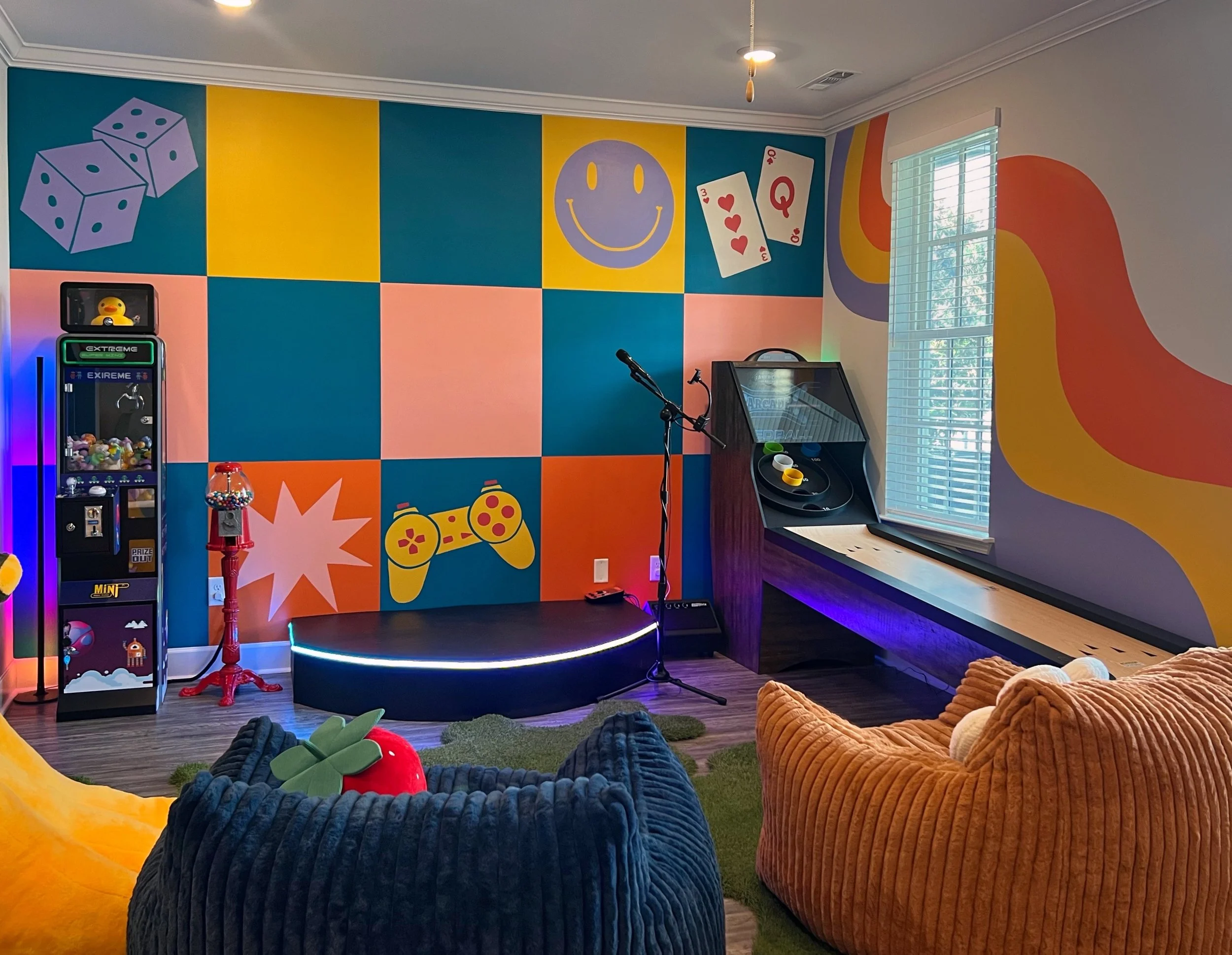 A colorful indoor game room featuring a painted wall with dice, a smiley face, playing cards, a game controller, and a pinball machine. There are plush bean bag chairs and stuffed toys on the floor.