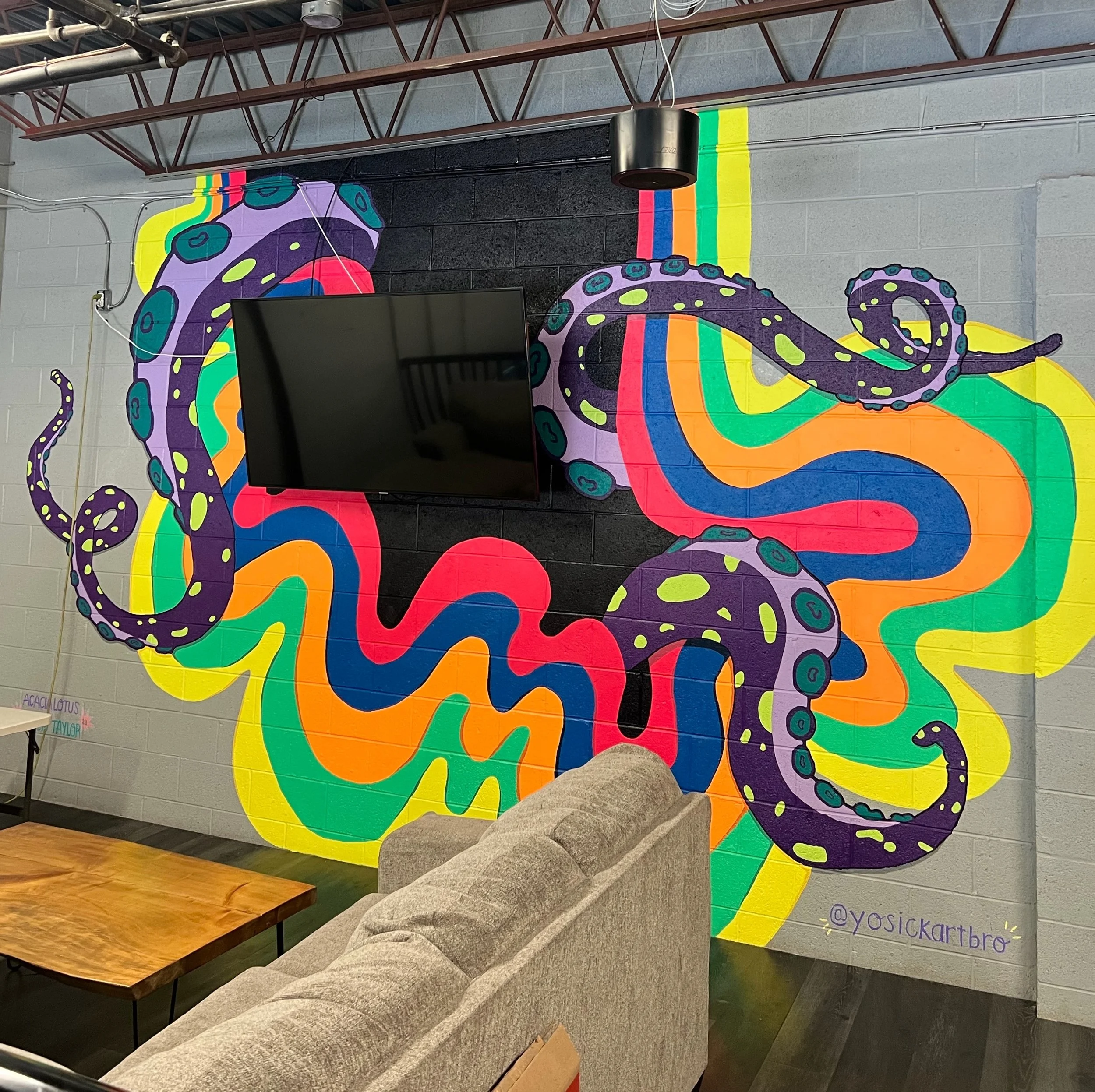 A colorful mural of an octopus painted on a cinder block wall in an indoor space. The octopus has purple, black, and yellow coloring with rainbow stripes in the background. There is a flat screen TV mounted on the wall, and a gray padded bench part i