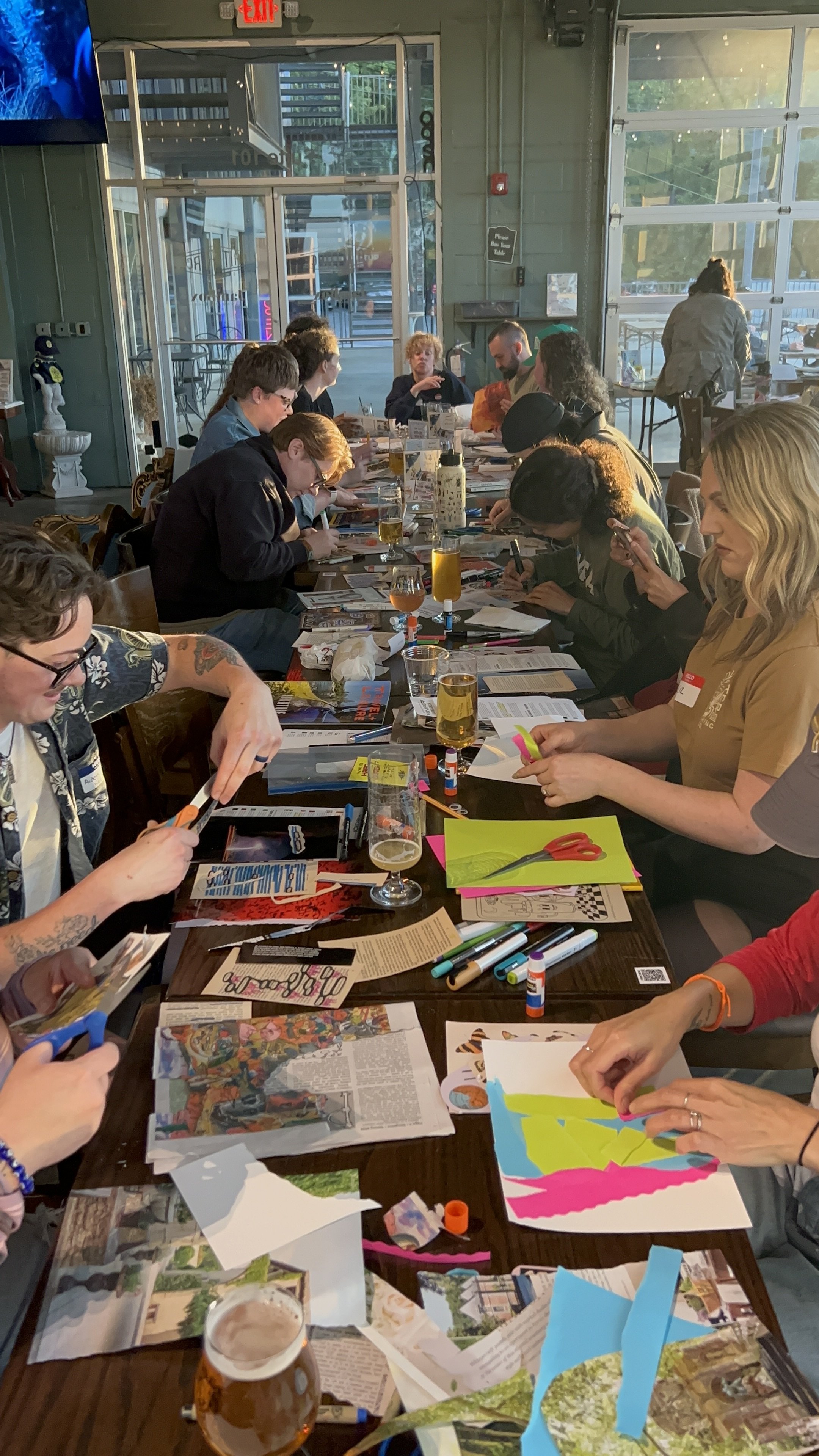 Group of people sitting around a long table engaged in a craft or art activity, with art supplies, papers, magazines, and drinks on the table inside a well-lit brewery at Nashville's Sketchbook Club hosted by Acacia Barron and Tory Lava