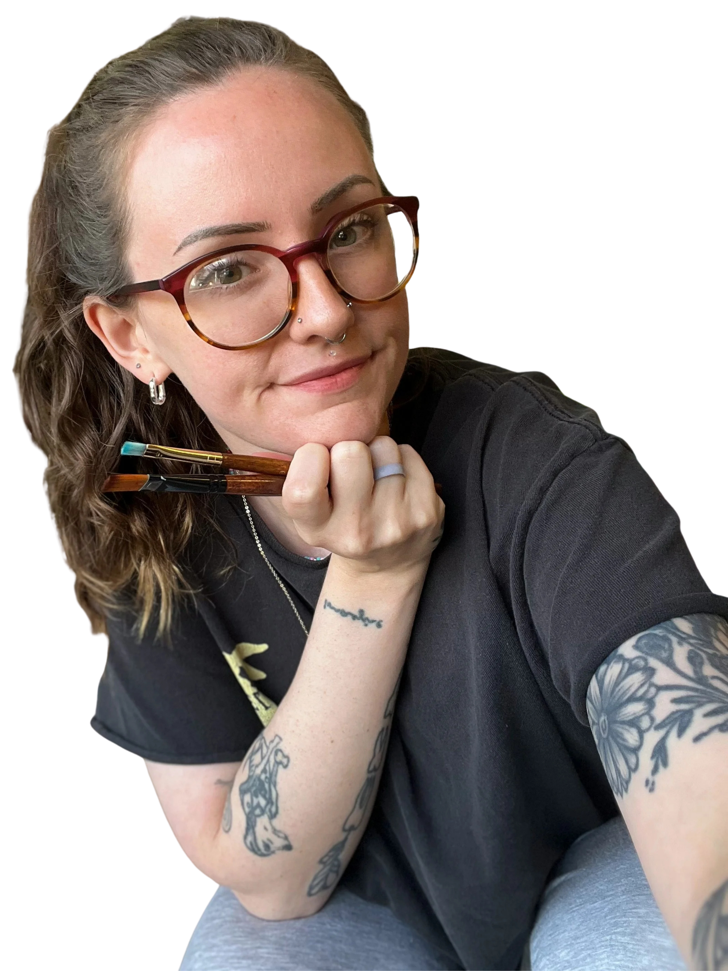 Close-up of a woman with light skin, curly hair, wearing glasses, and tattoos on her arms, sitting with her chin resting on her hand and smiling at the camera.