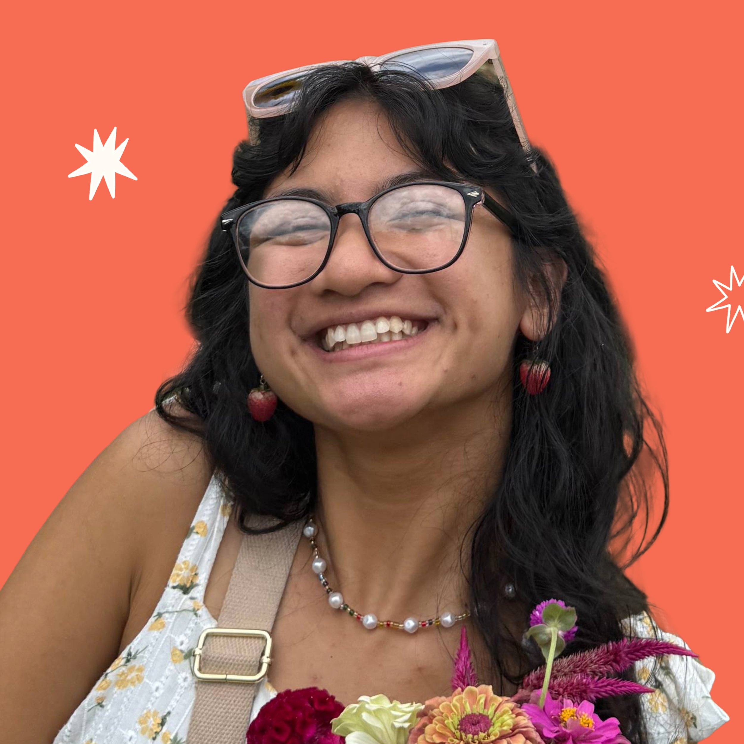 Close-up of a smiling woman with dark curly hair, wearing glasses, jewelry, a floral dress, and holding colorful flowers against a bright orange background with white and black starburst decorations.
