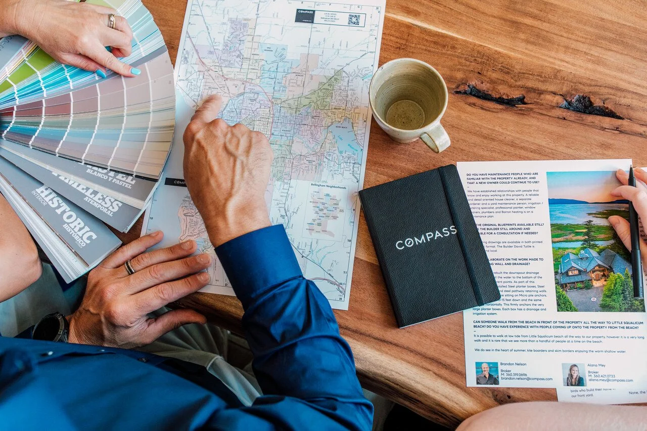 A tabletop scene with hands pointing at a map alongside color swatches, a notebook labeled 'COMPASS,' a cup, and a real estate brochure.
