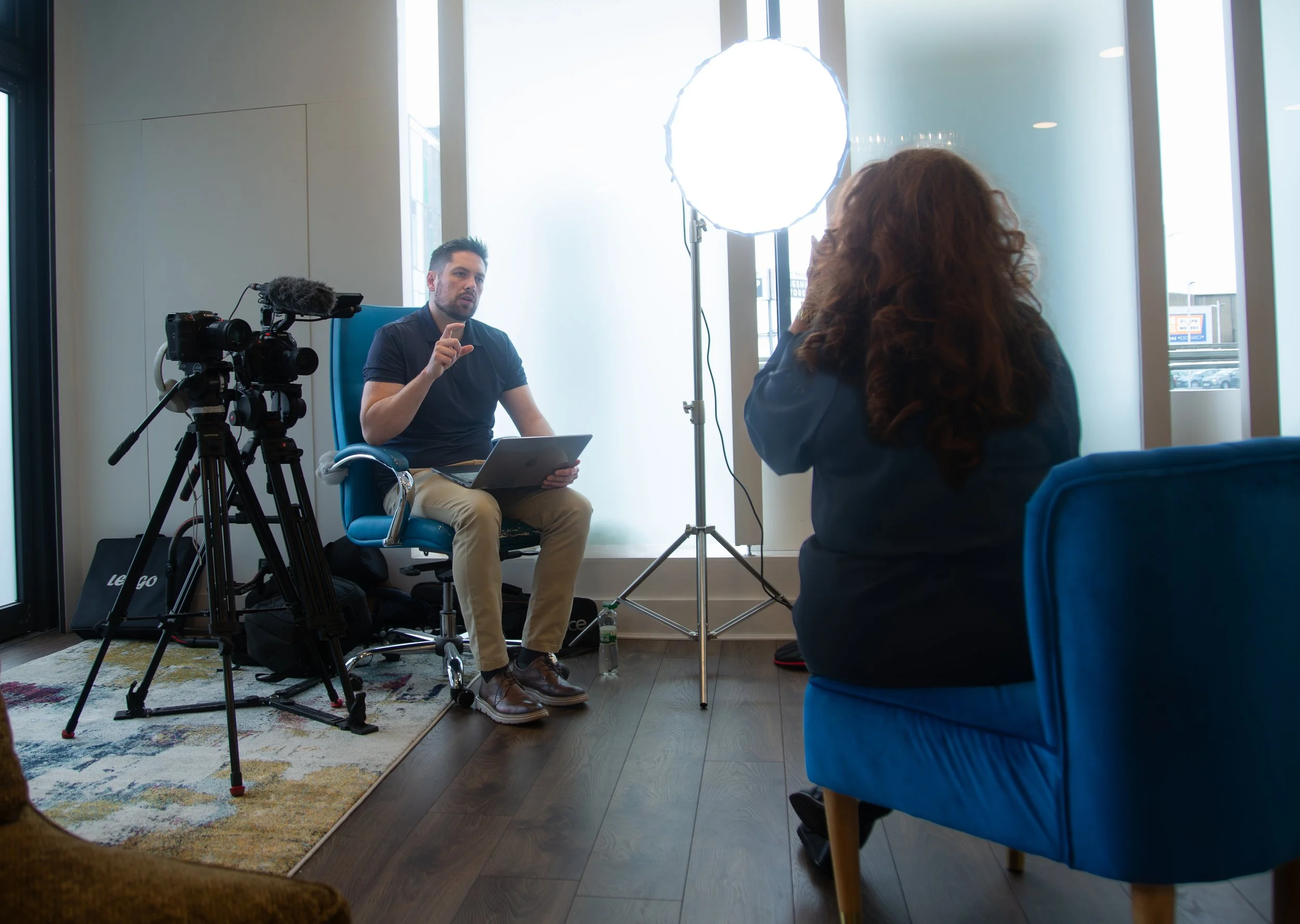 A man being interviewed indoors with a camera and a bright light on a stand in front of him, and a woman standing with her back to the camera, facing the man.
