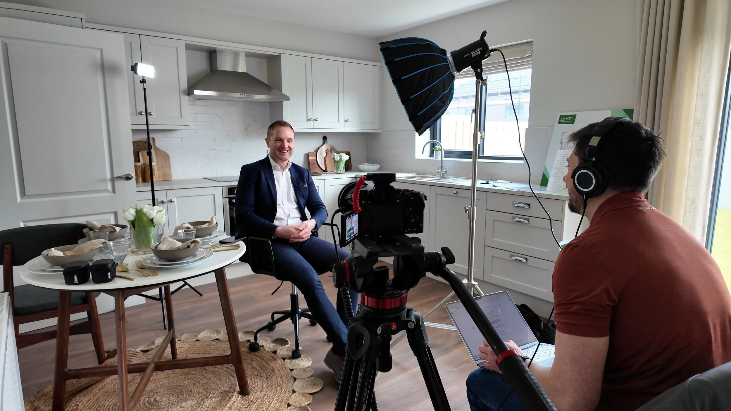 A man in a suit sitting at a dining table being filmed by a camera crew in a kitchen setting.