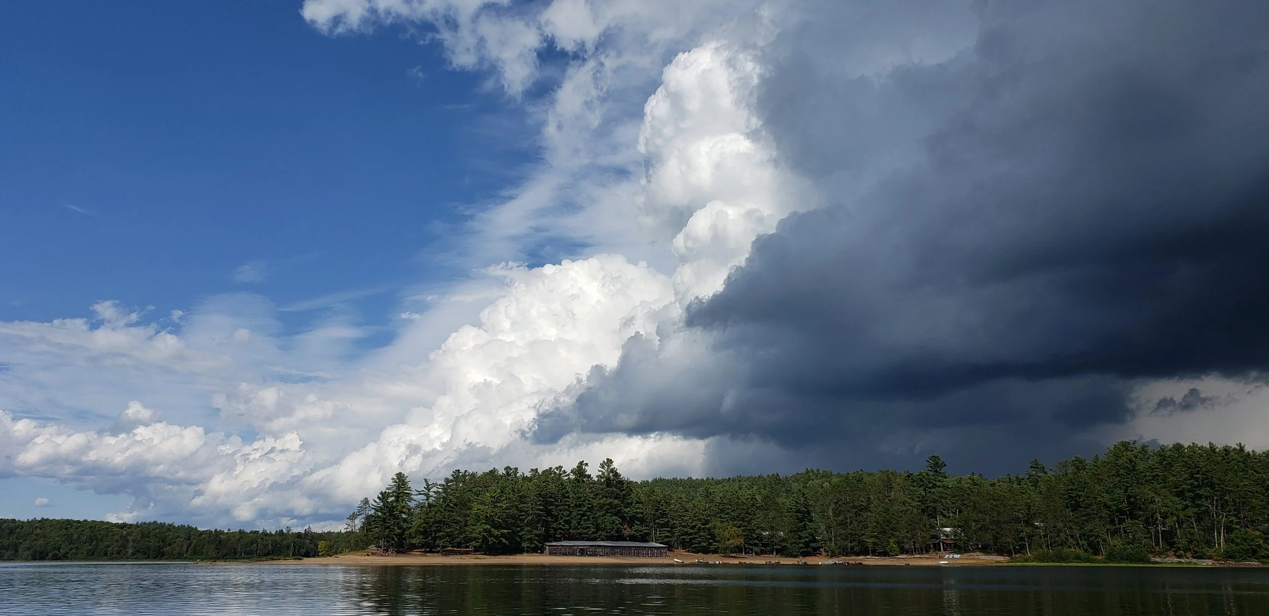 A large lake with a forested shoreline under a dramatic sky with dark storm clouds and scattered white clouds.