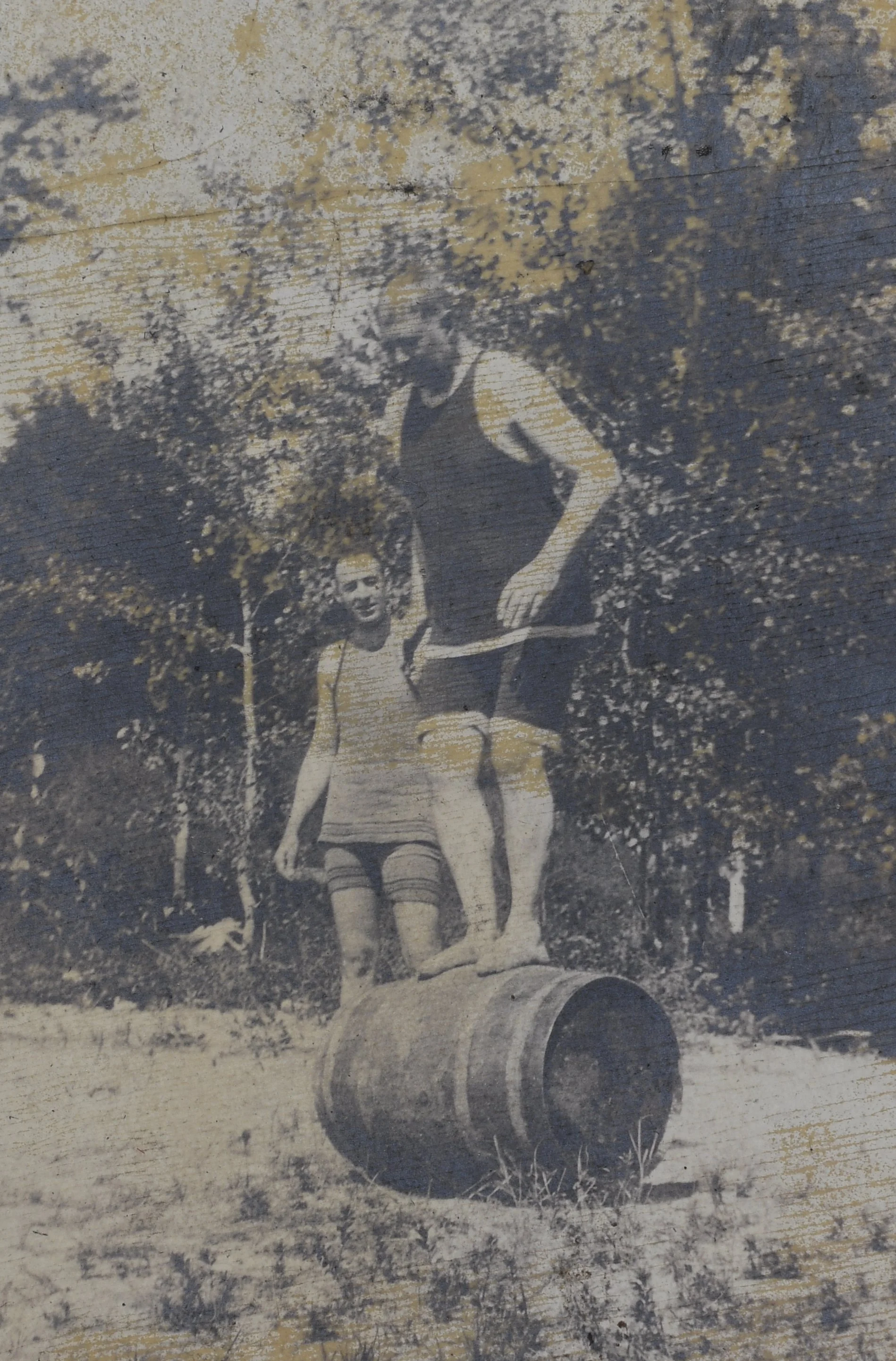 A vintage black-and-white photo of an outdoor scene with a woman standing on a barrel, holding hands with a person who is balancing on top of the barrel, surrounded by trees and foliage.