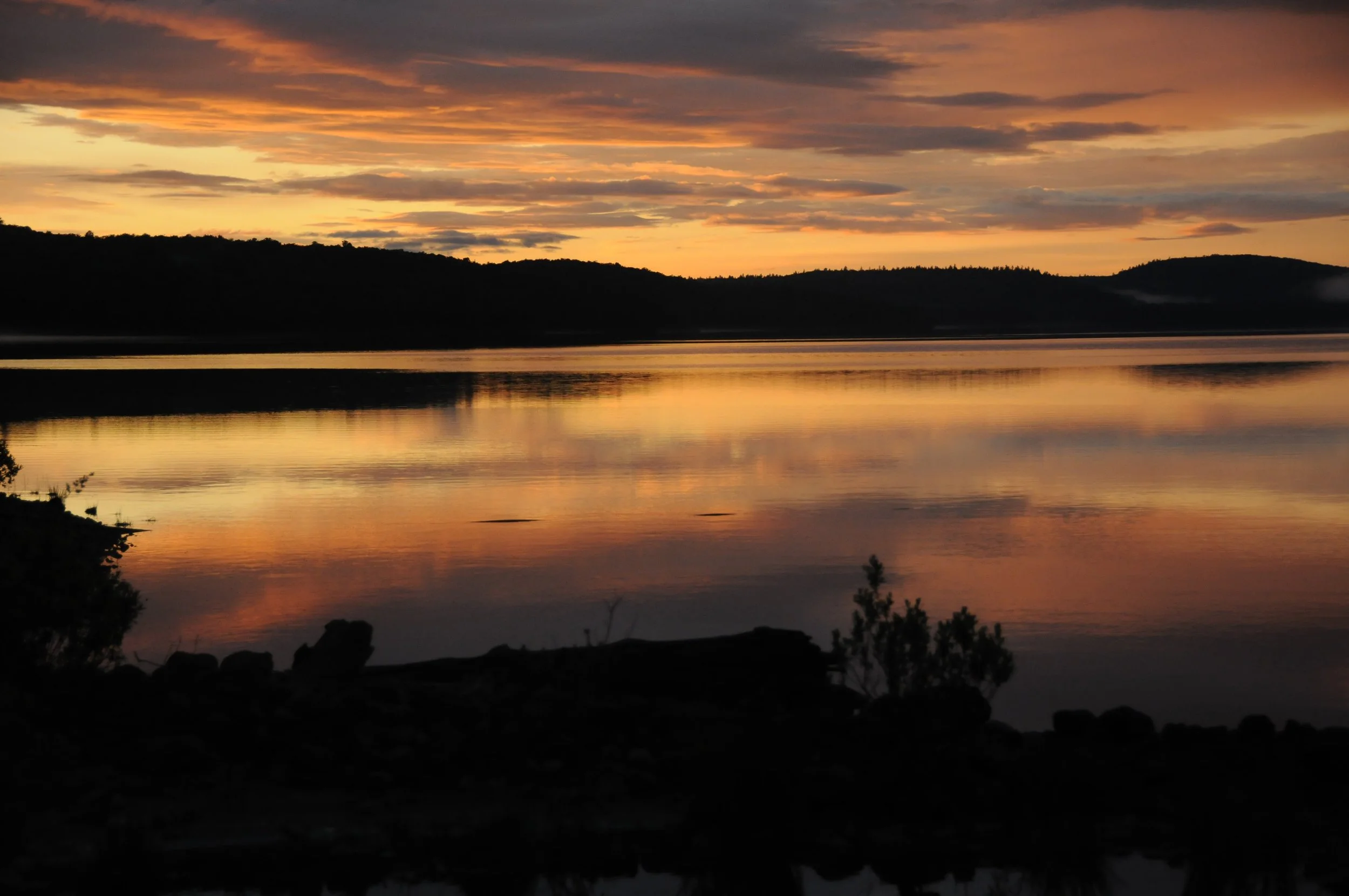 A sunset over a calm lake with orange, pink, and purple hues in the sky, reflecting on the water, with dark silhouettes of hills and bushes in the foreground.