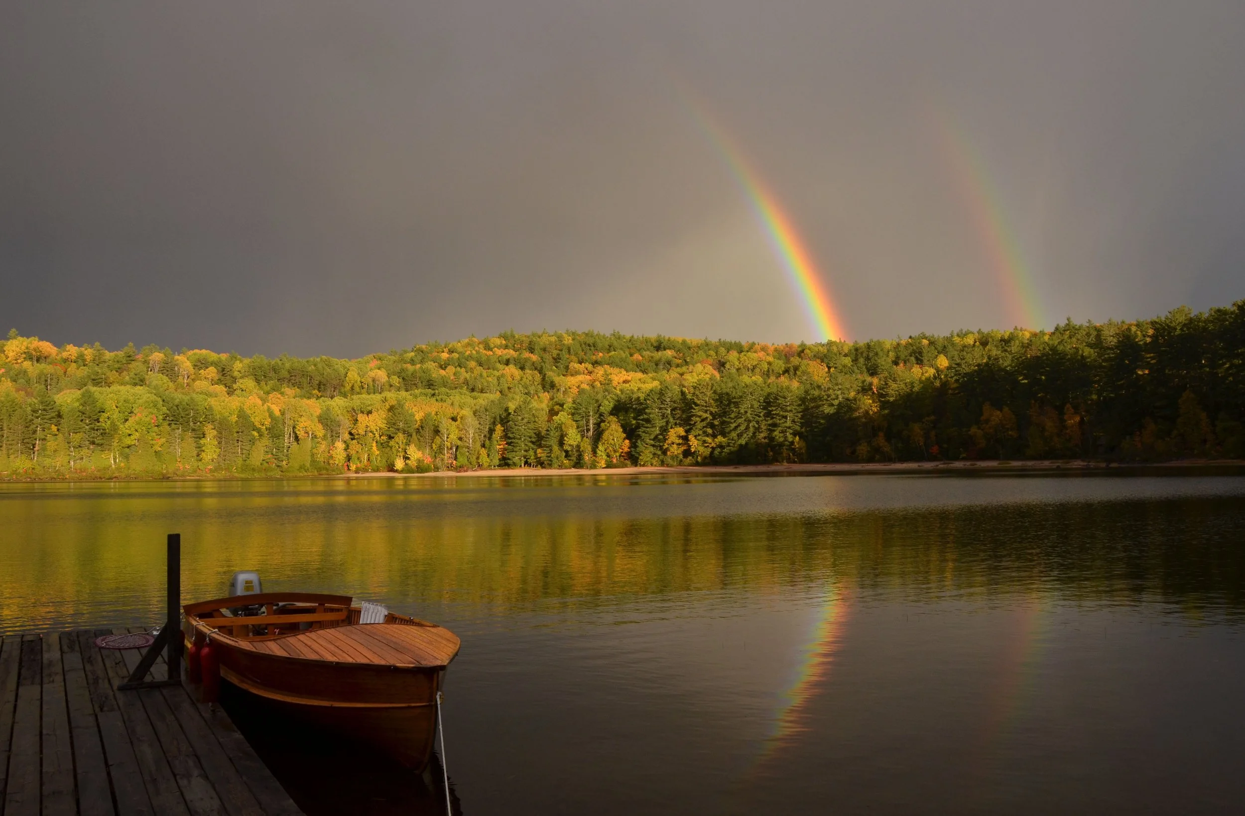 A wooden boat docked at a lake with a double rainbow arcing across a dark and stormy sky, and a forested shoreline in the background.