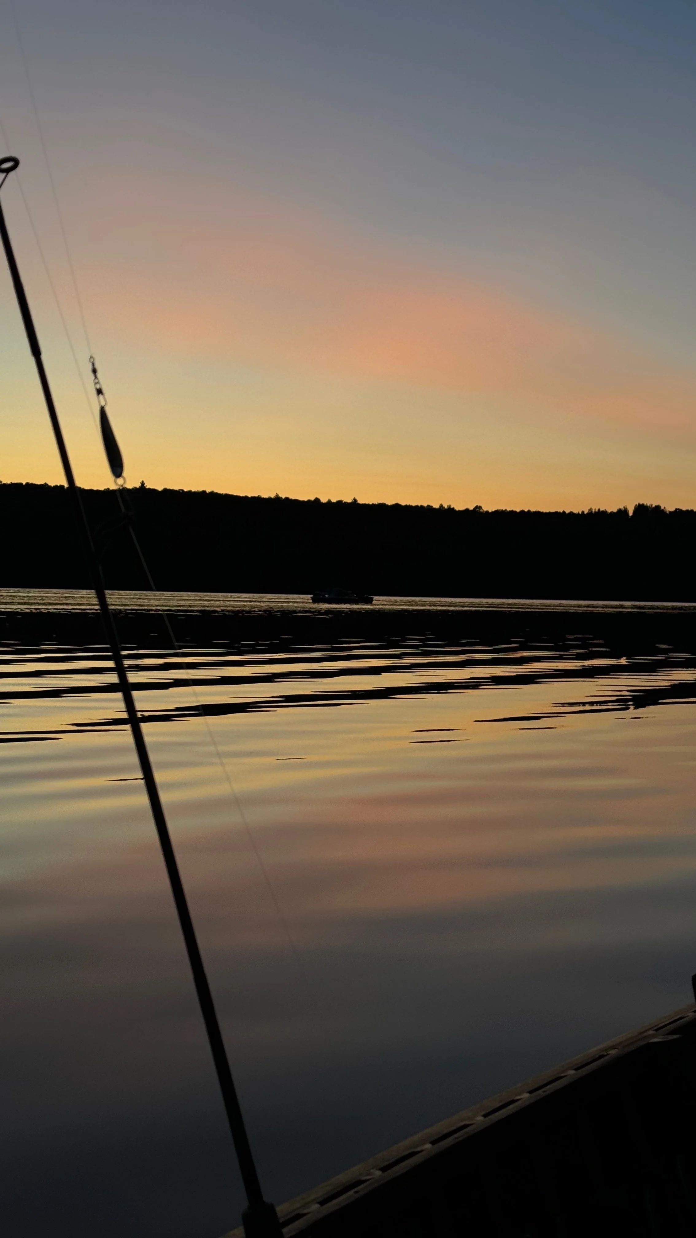 A serene lake scene at sunset or sunrise, with calm water reflecting the sky's pastel colors, a distant boat on the water, and a boat's fishing rod in the foreground.