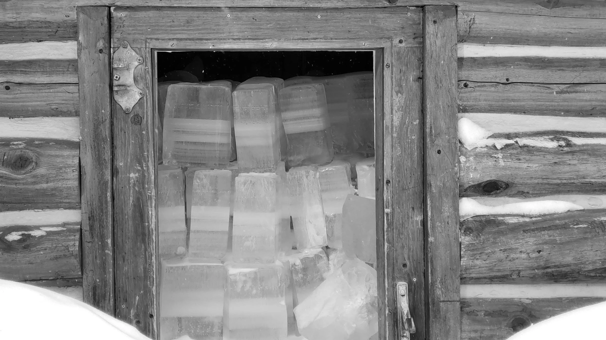 A small wooden cabin wall with a window showcasing stacked ice blocks inside, with snow on the outside.
