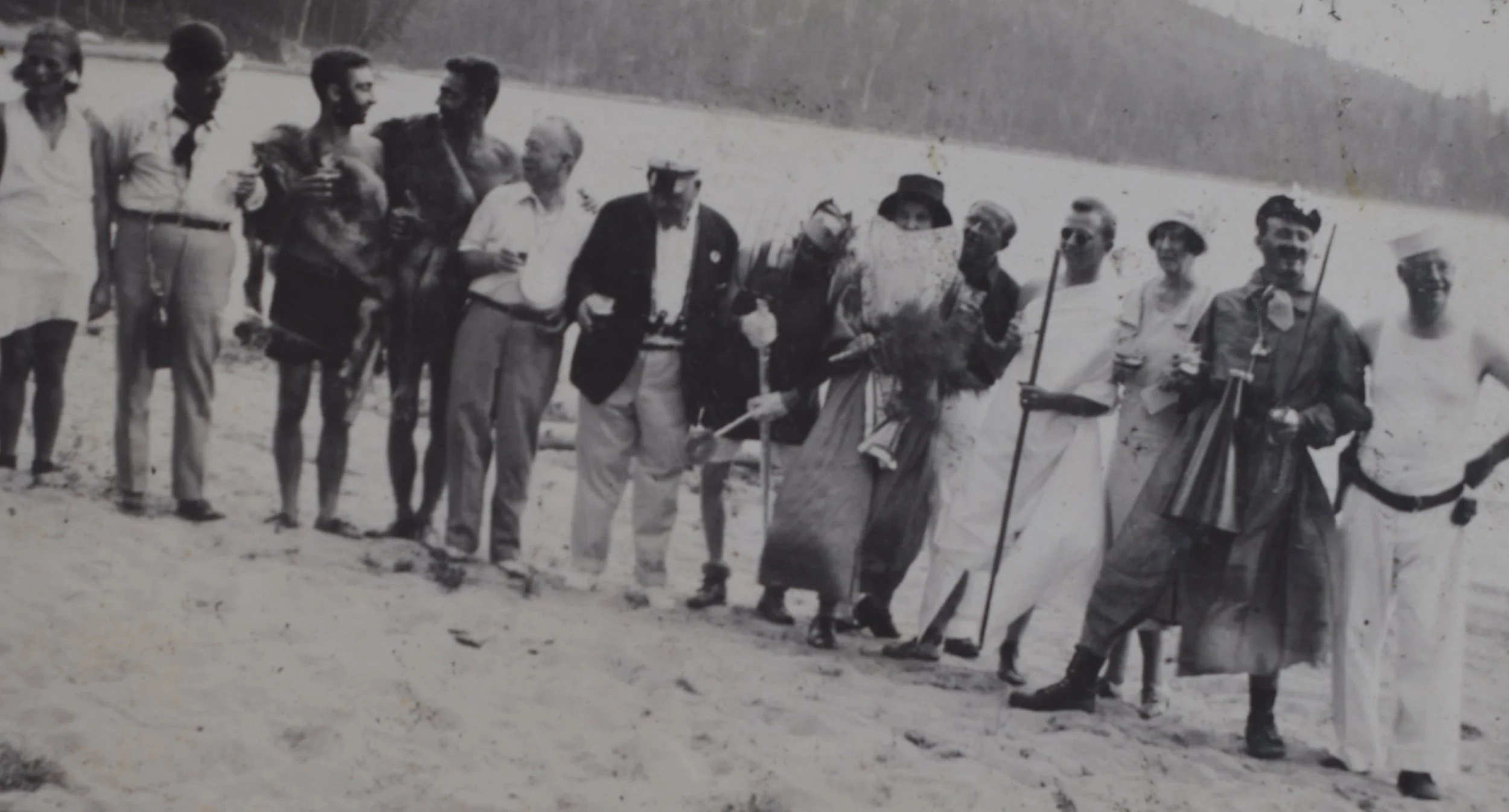 Black and white photograph of a group of people standing on the beach, some dressed in traditional attire and others in casual clothing, smiling and standing close together.