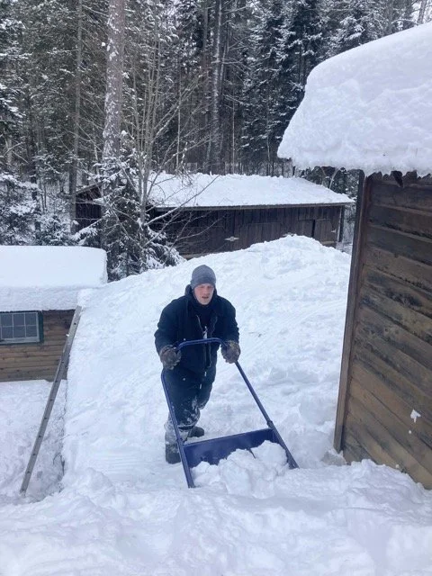 Person wearing winter clothing using a snow shovel to clear snow from a pathway outdoors surrounded by wooden structures and snow-covered trees.