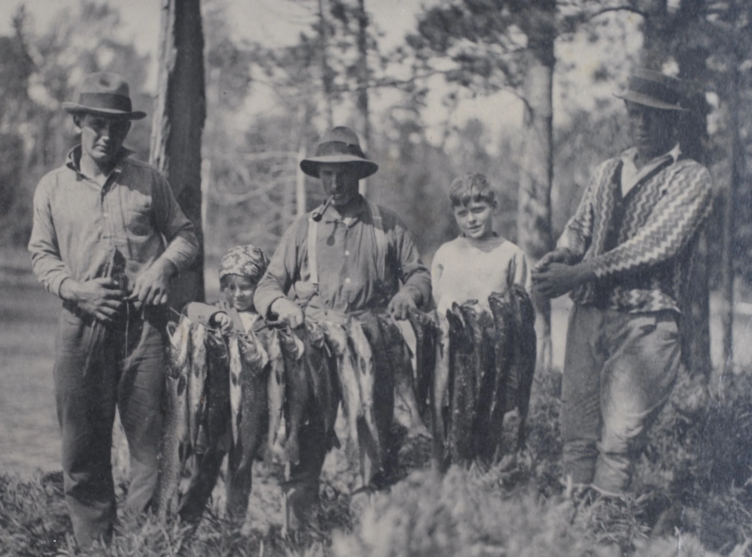 Black and white photo of five fishermen standing outdoors in a forested area, holding large fish they caught.