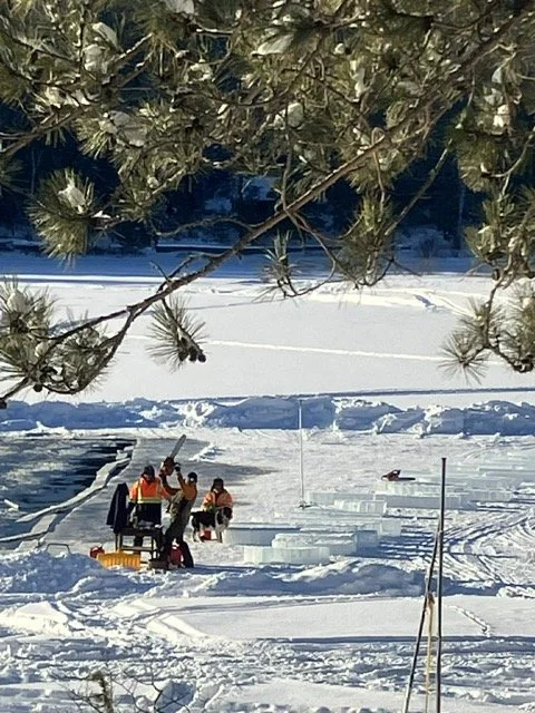 A group of people and equipment on a snow-covered surface near a frozen lake, with trees and a forested background, under a clear sky.