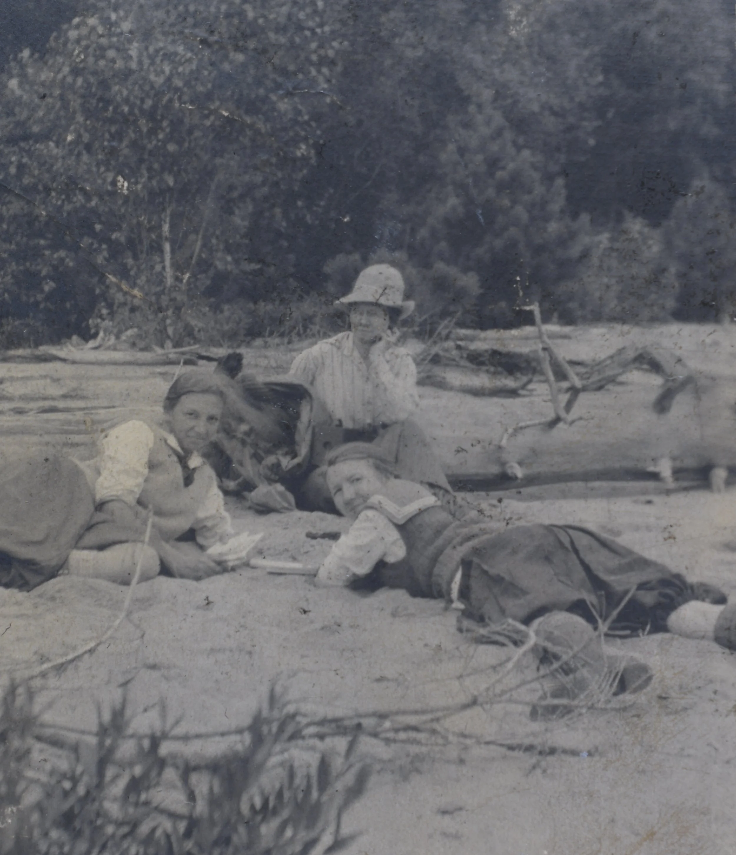 Three women are lying on the ground and relaxing outdoors, with a large log or fallen tree behind them and a forested background.