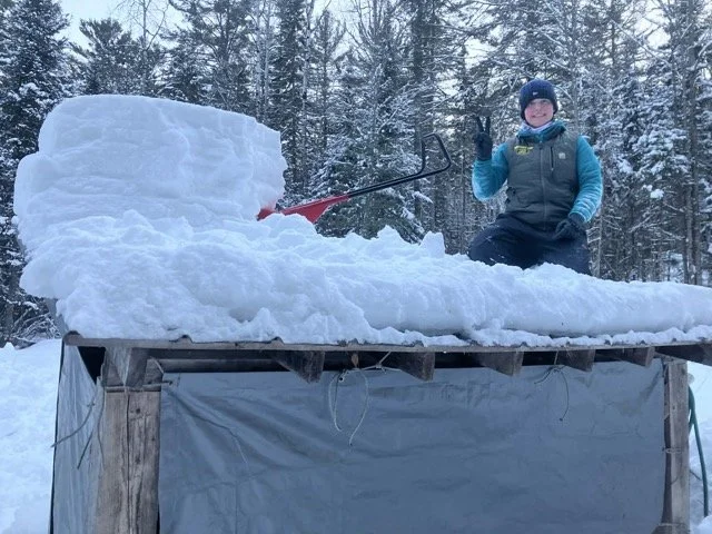 A person in winter clothing sitting on a snow-covered platform in a snowy forest, smiling and waving, with a large snow sculpture and a snow shovel nearby.