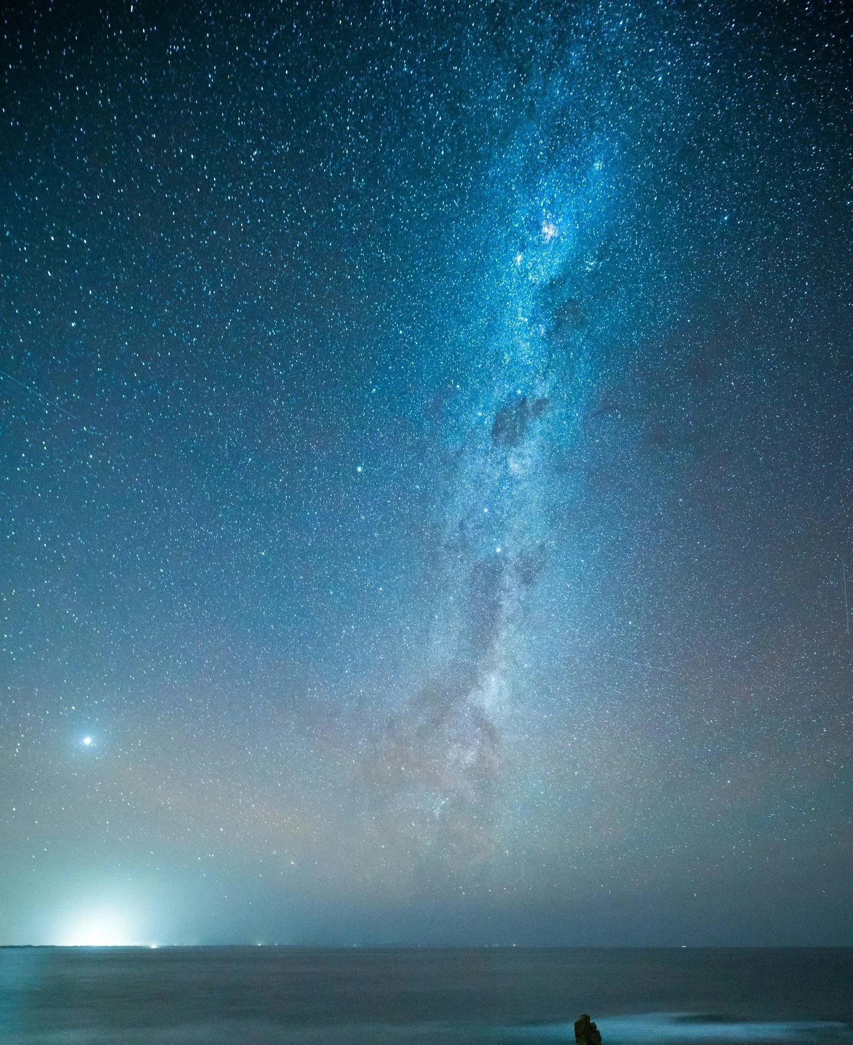 Starry night sky filled with countless stars above a calm ocean, with a silhouette of a person sitting on a rock at the shoreline.