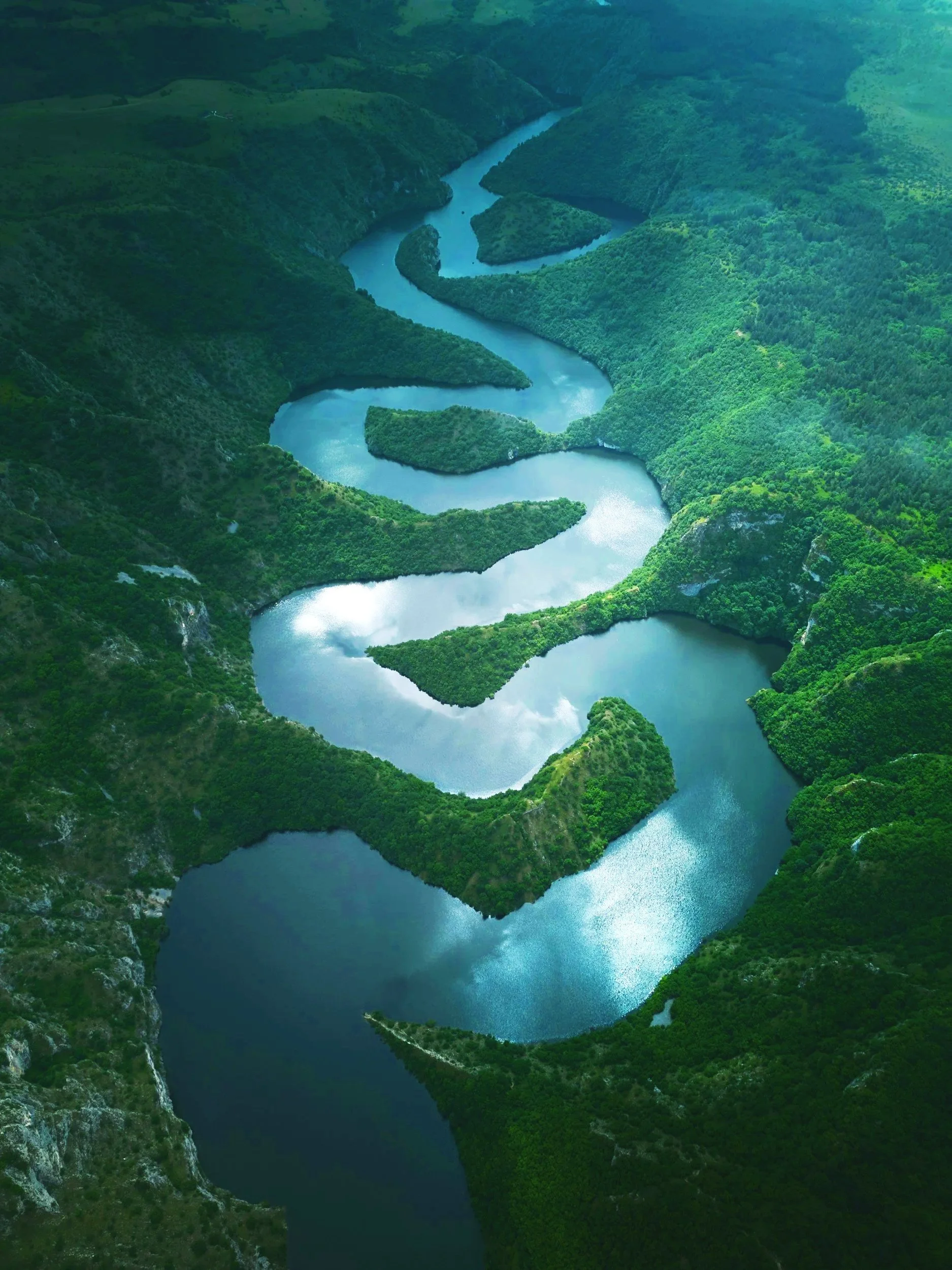 Aerial view of a winding turquoise reservoir surrounded by lush green, forested hills and rocky cliffs