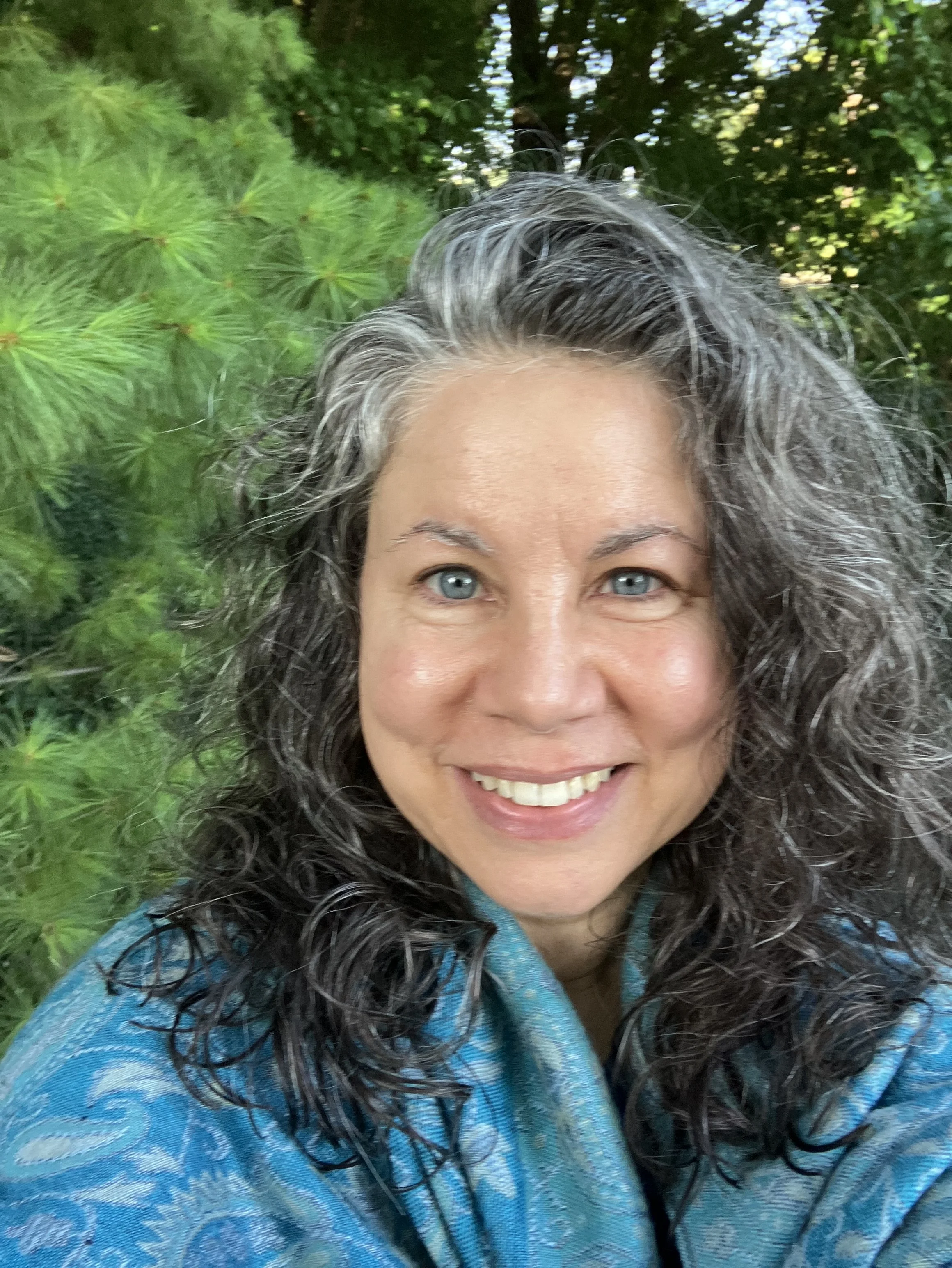 A woman with long, curly gray and black hair, smiling outdoors with a background of green foliage and trees.