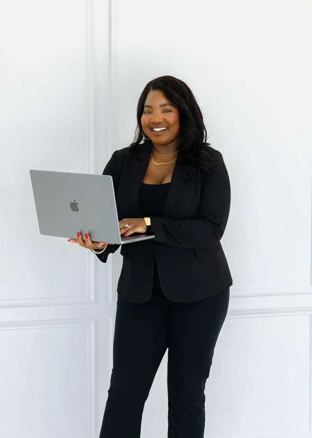 Eripitan Osagie-Sule, Founder of Light On A Hill Marketing is a woman in a black blazer holding a silver MacBook, smiling, standing against a white wall.