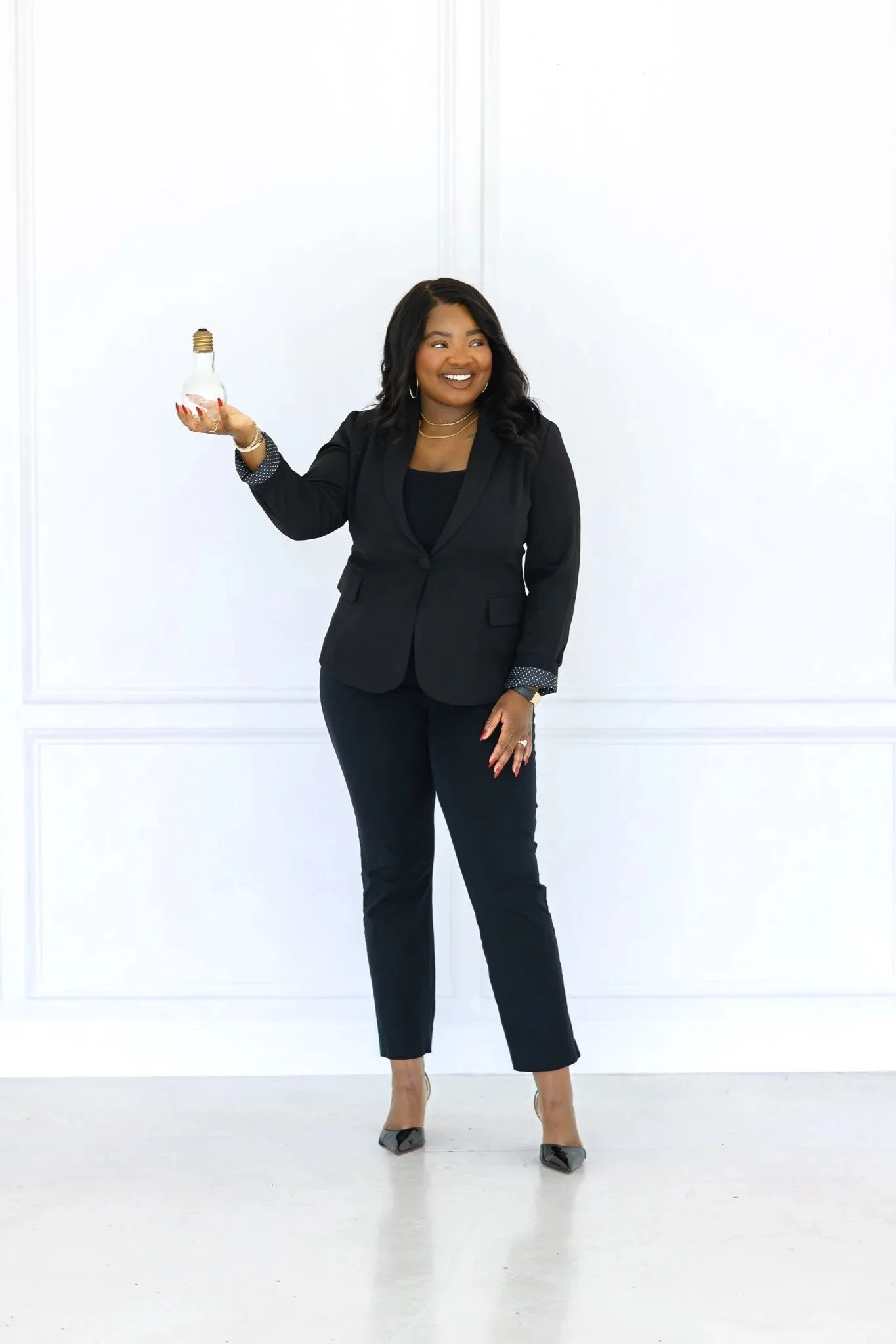 Eripitan Osagie-Sule, Founder of Light On A Hill Marketing is a  woman in a black business suit holding a lightbulb in her right hand, smiling and standing in front of a plain white wall.