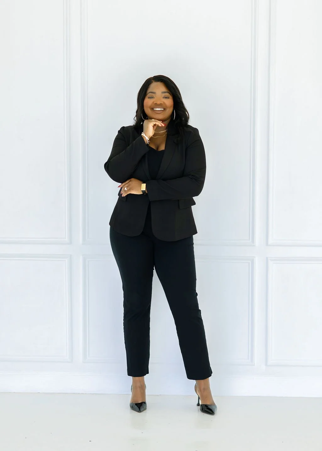 Eripitan Osagie-Sule, Founder of Light On A Hill Marketing with black hair dressed in a black suit standing against a white wall, smiling, with one hand on her chin and the other on her waist.
