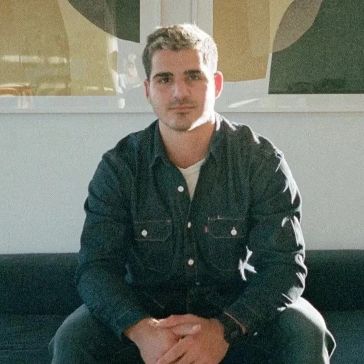 A young man with short, dark hair, wearing a dark denim jacket over a white shirt, sitting on a black bench or sofa in a well-lit indoor setting.
