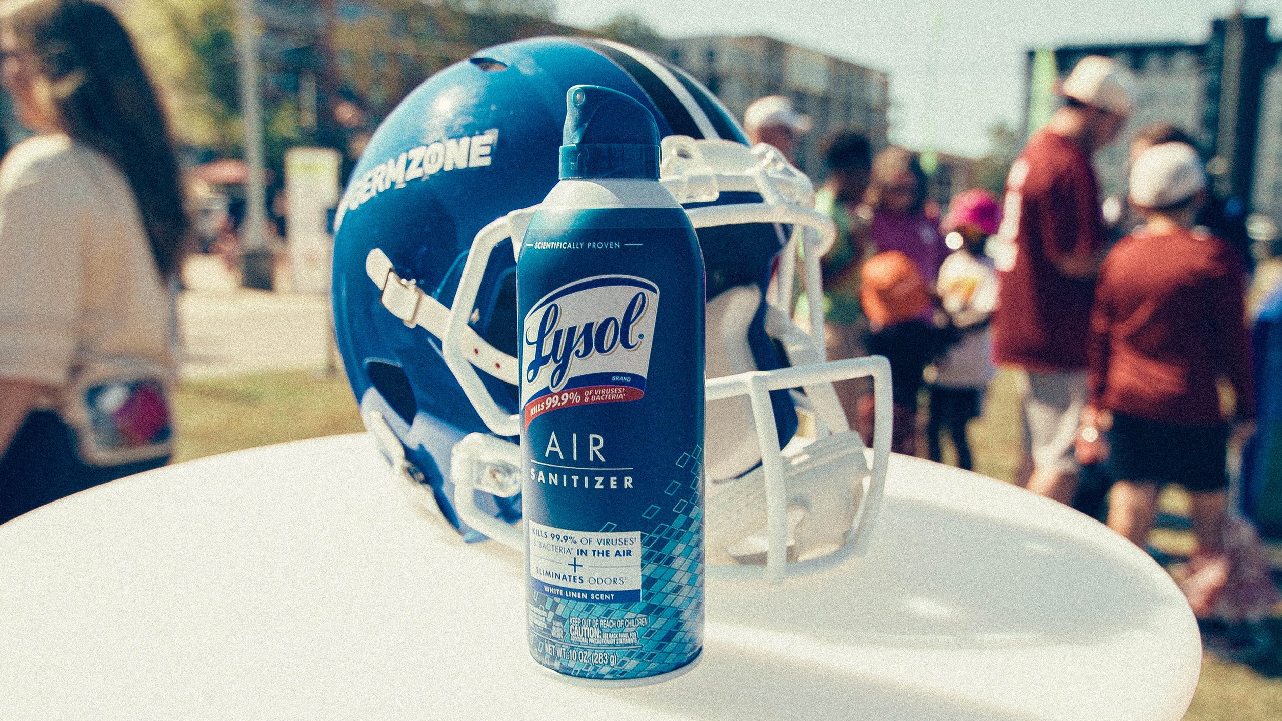 A bottle of Lysol air sanitizer placed on top of a white surface, with a blue football helmet featuring the word "HORIZON" in the background and a group of people in casual attire at an outdoor event.