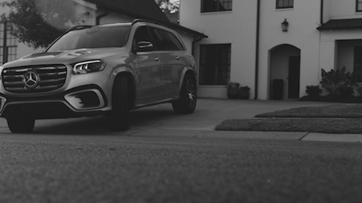 A silver Mercedes-Benz SUV parked in front of a house with a lawn and sidewalk in the suburban neighborhood.