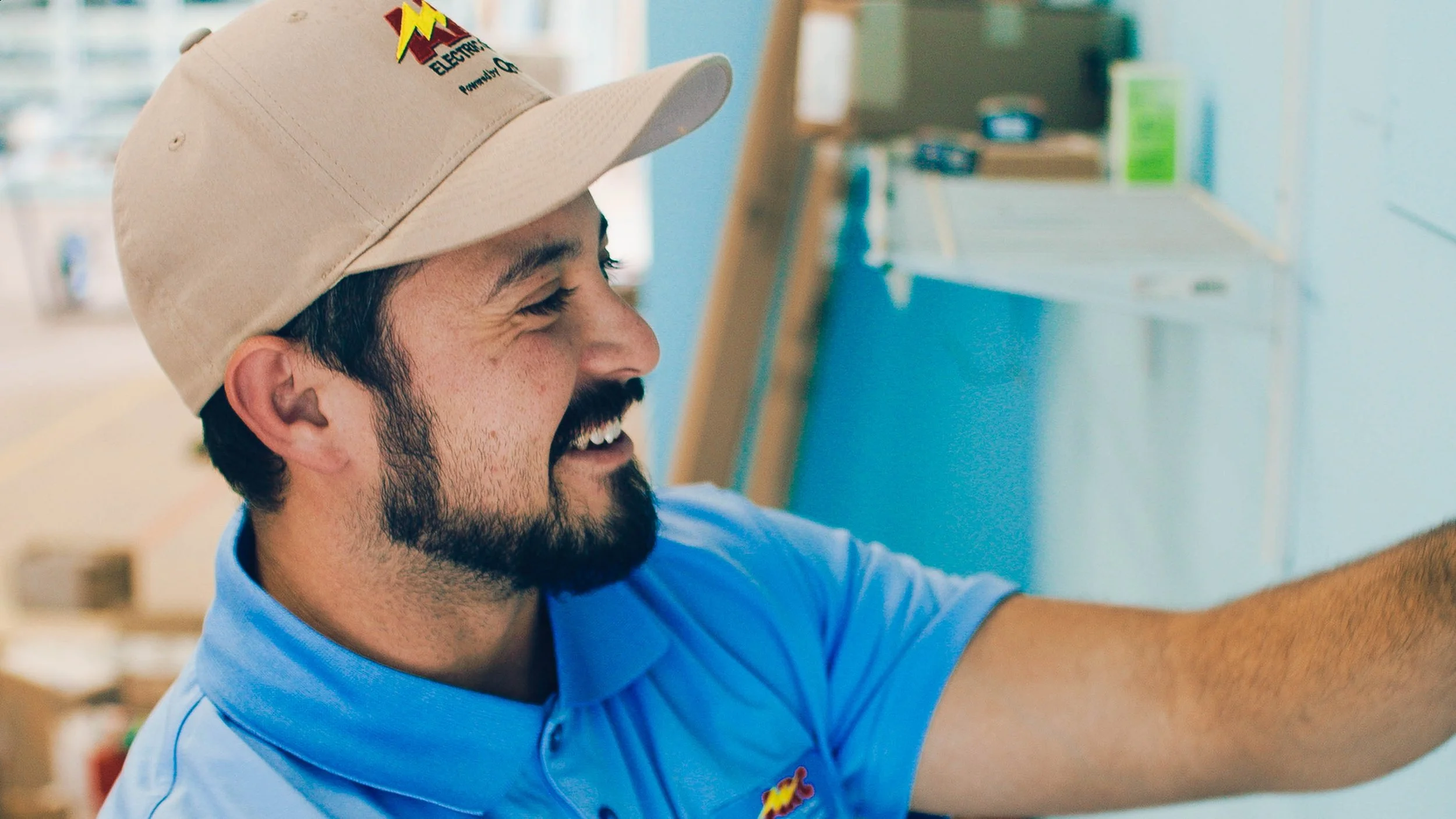 A man smiling, wearing a beige baseball cap with an electrician logo and a light blue polo shirt, working in a room with a light blue wall and shelves in the background.