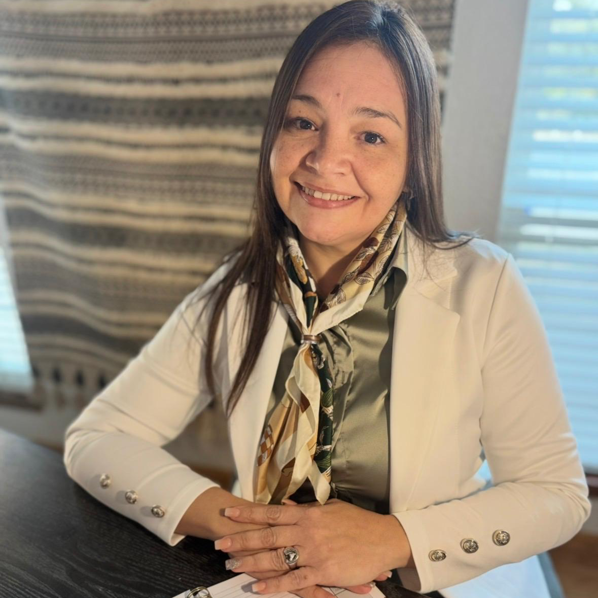 Montessori Guide & Prepared Adult Instructor, Monica Portillo, with long brown hair smiling, wearing a beige blazer with decorative buttons, a scarf around her neck, and a ring on her finger, sitting at a table with her hands clasped.