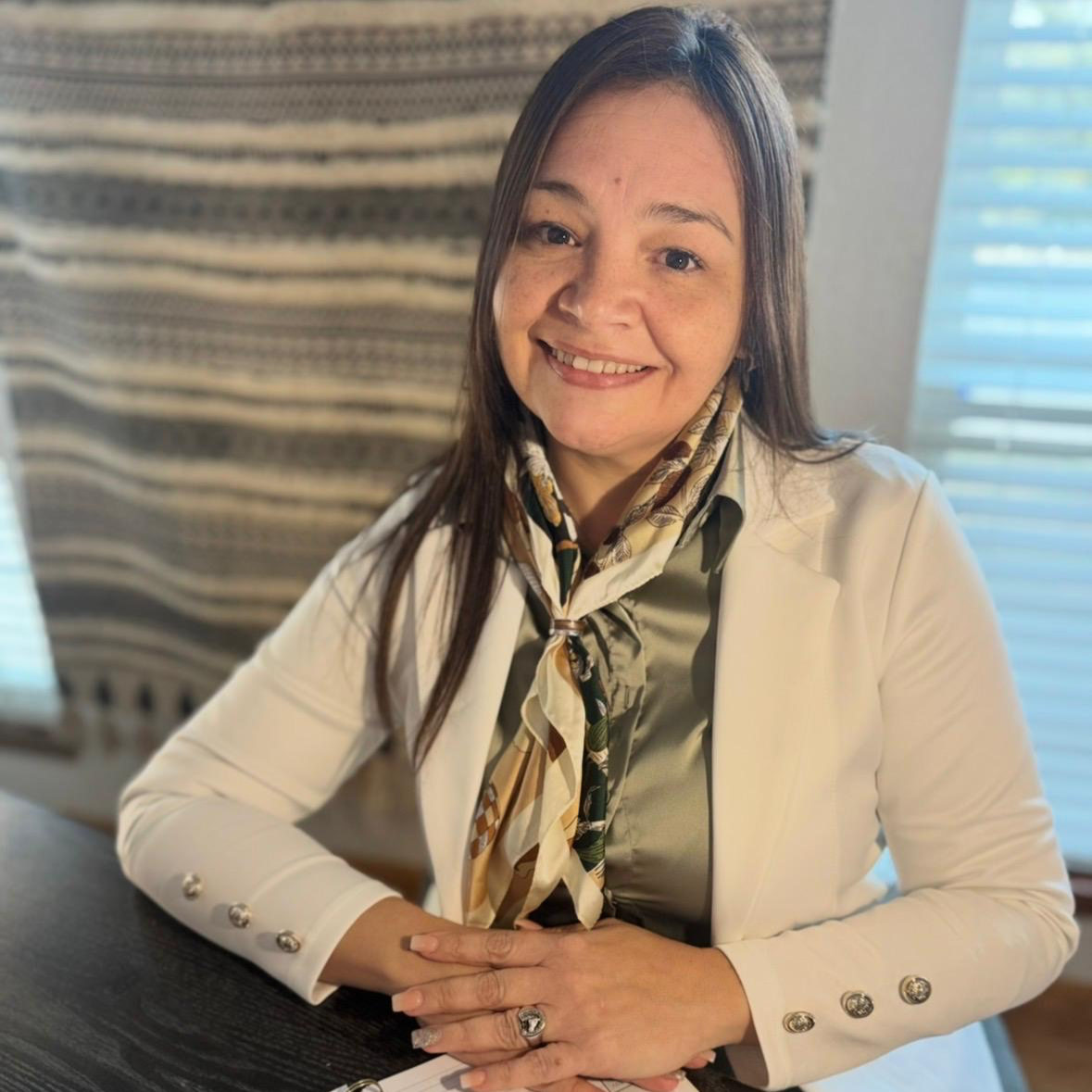 Montessori Guide & Prepared Adult Instructor, Monica Portillo, with brown hair, wearing a beige blazer and a colorful scarf, sitting at a desk with her hands clasped, smiling at the camera indoors with window blinds in the background.