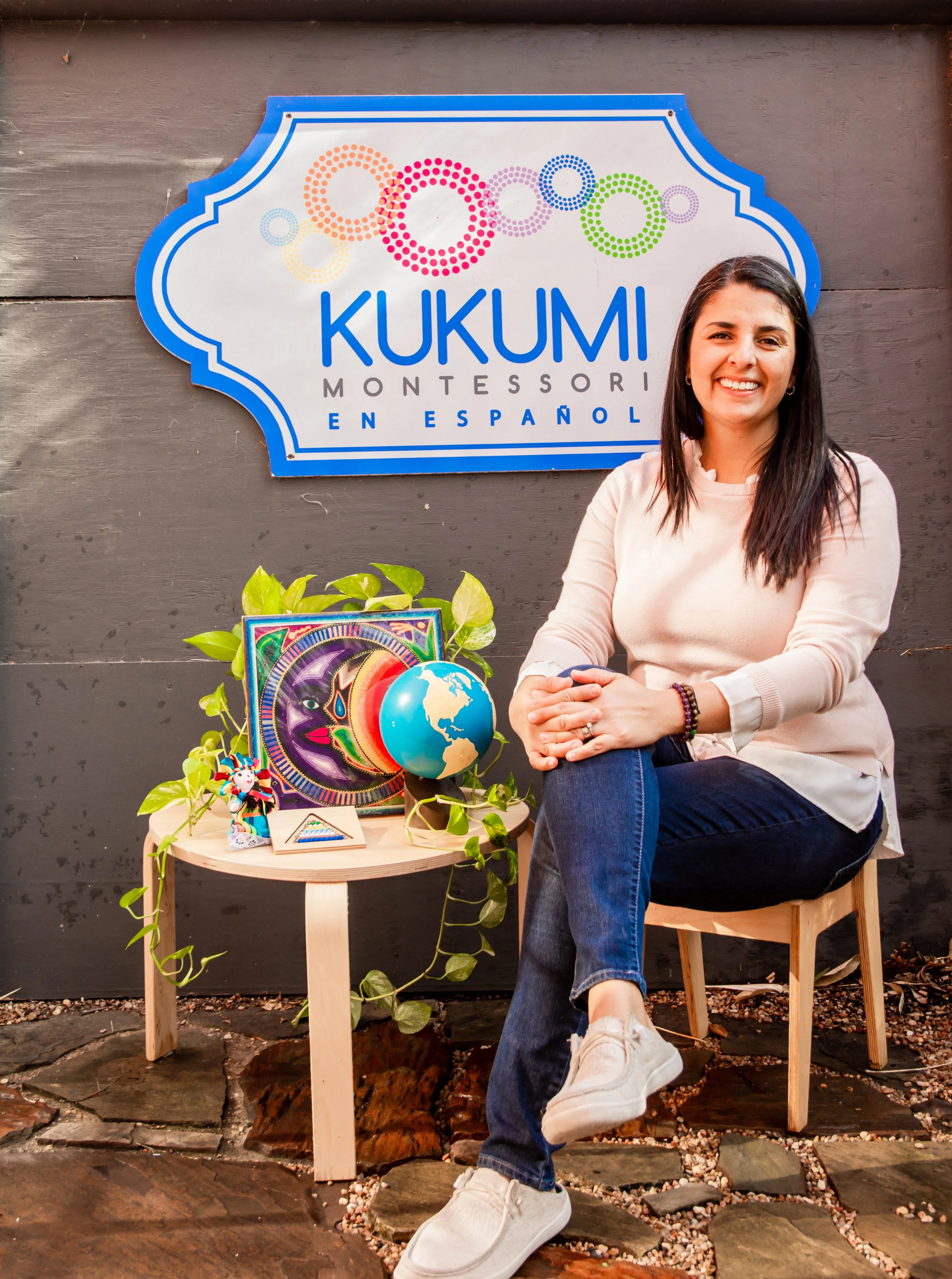 Head of Kukumi Mrs. Daniela Garcia sitting on a wooden chair smiling, with a small table beside her decorated with colorful items, in front of a large sign that reads 'Kukumi Montessori en Español.'