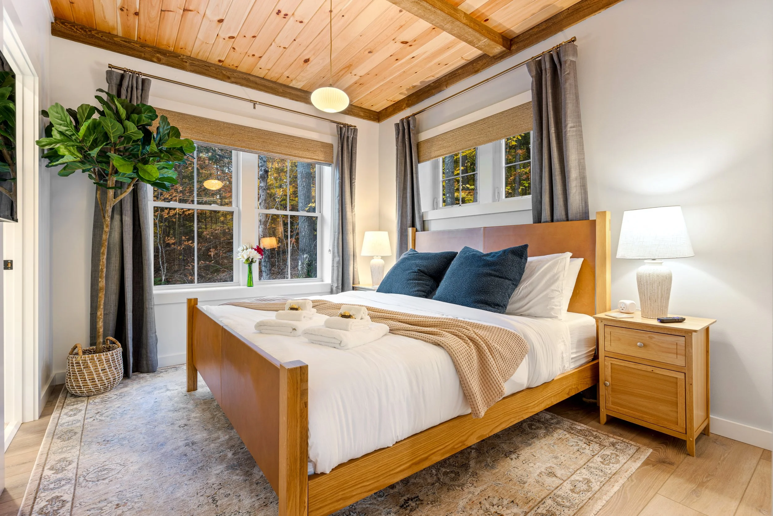 Cozy bedroom with large window showing trees outside, bed with white linens, beige blanket, and plaid pillows, and a green upholstered bench at foot of bed.