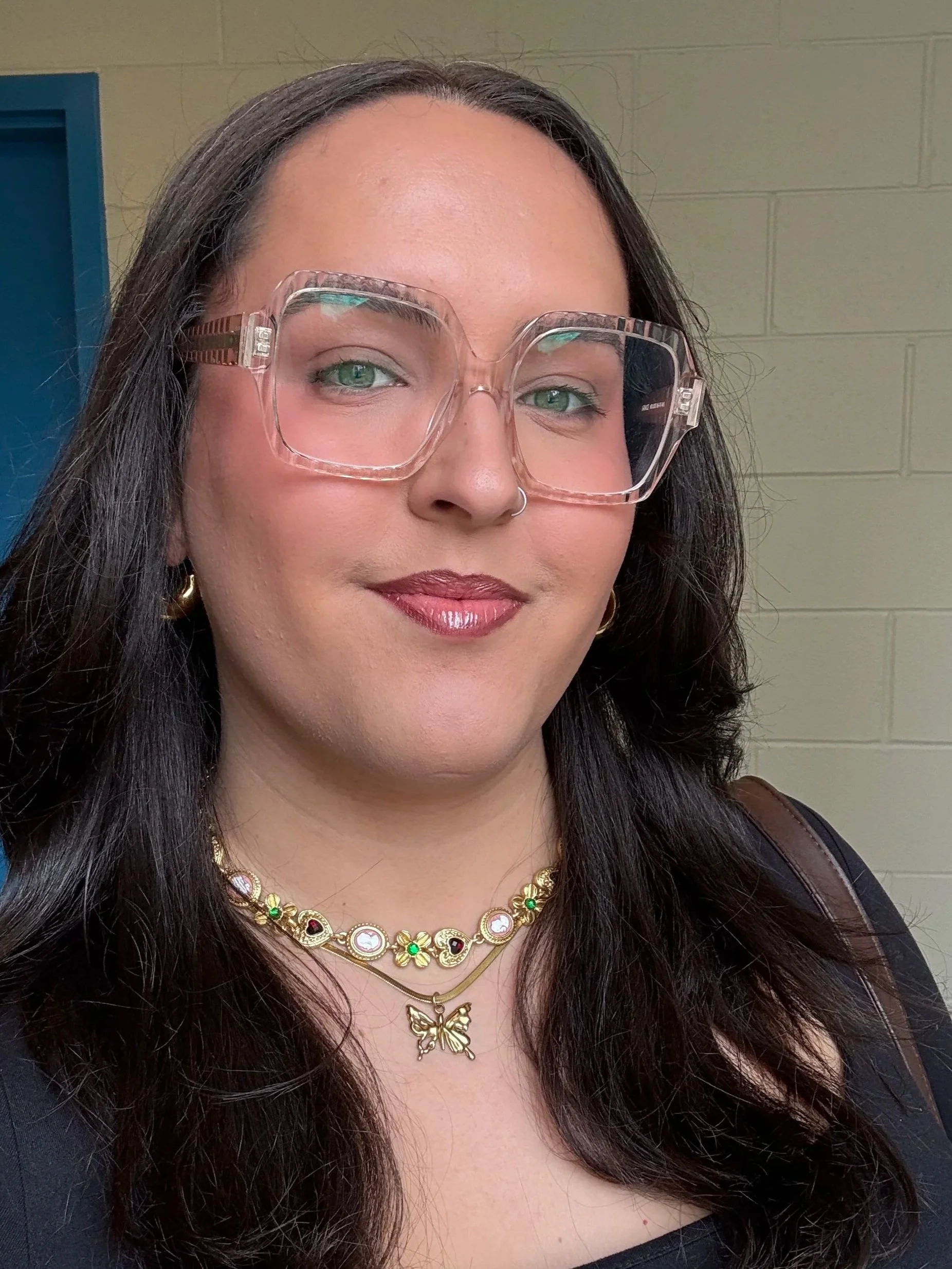 Une femme avec des cheveux longs porte des lunettes transparentes, un collier avec des pendentifs en or, et un autre collier doré avec un papillon, elle porte un maquillage discret et un sourire léger, contre un mur beige avec une porte bleue à gauche.